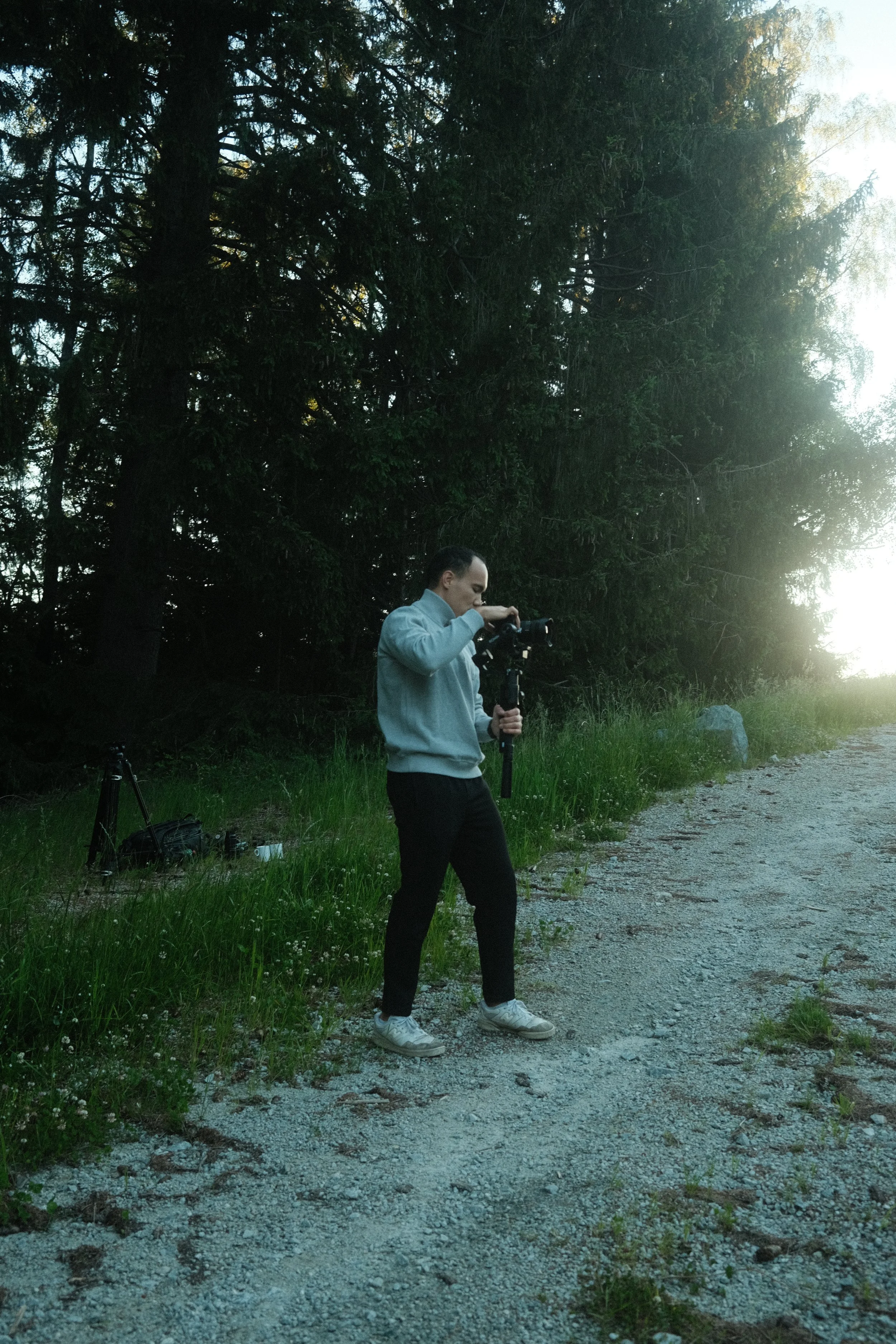 Junge Person steht im Freien auf einem Kiesweg, hält einen Gimbal mit Kamera, im Hintergrund Bäume und Sonnenlicht.