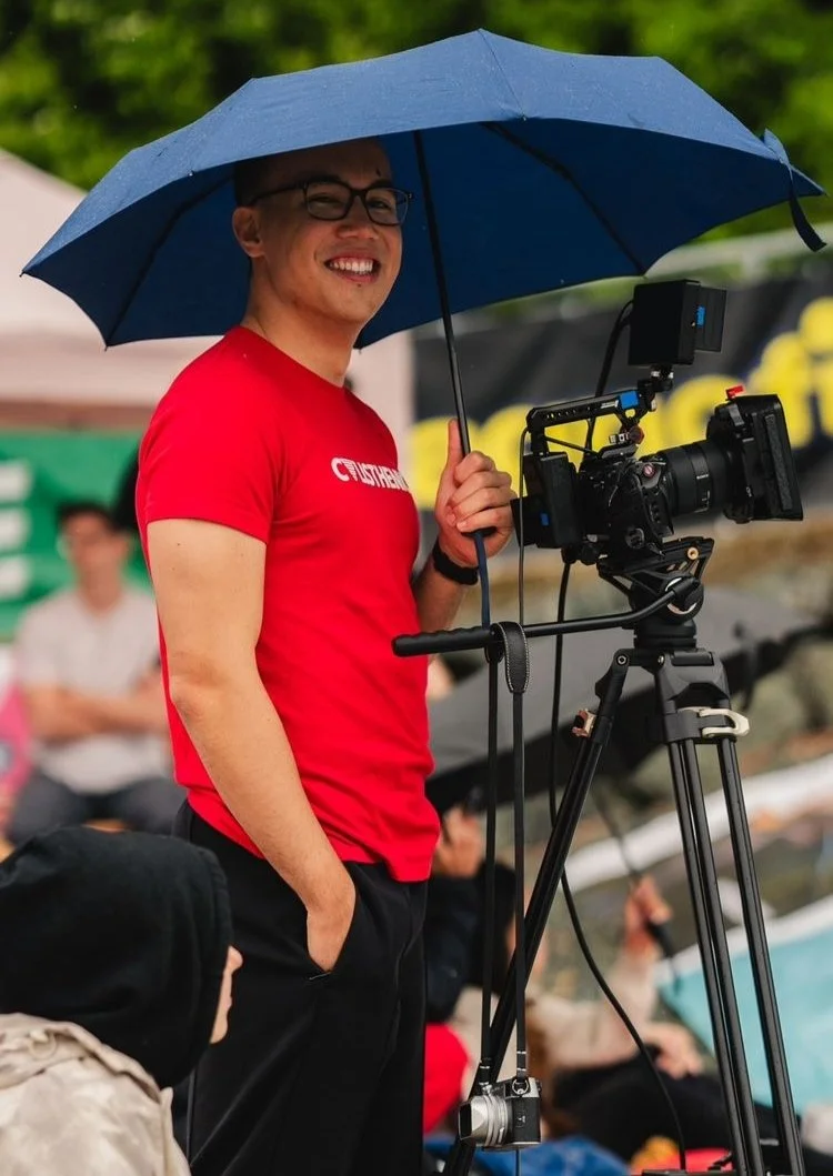 Ein Mann mit Brille und rotem T-Shirt steht bei einem Outdoor-Event hinter einer Kamera auf einem Stativ und hält einen blauen Regenschirm, während er lächelt.