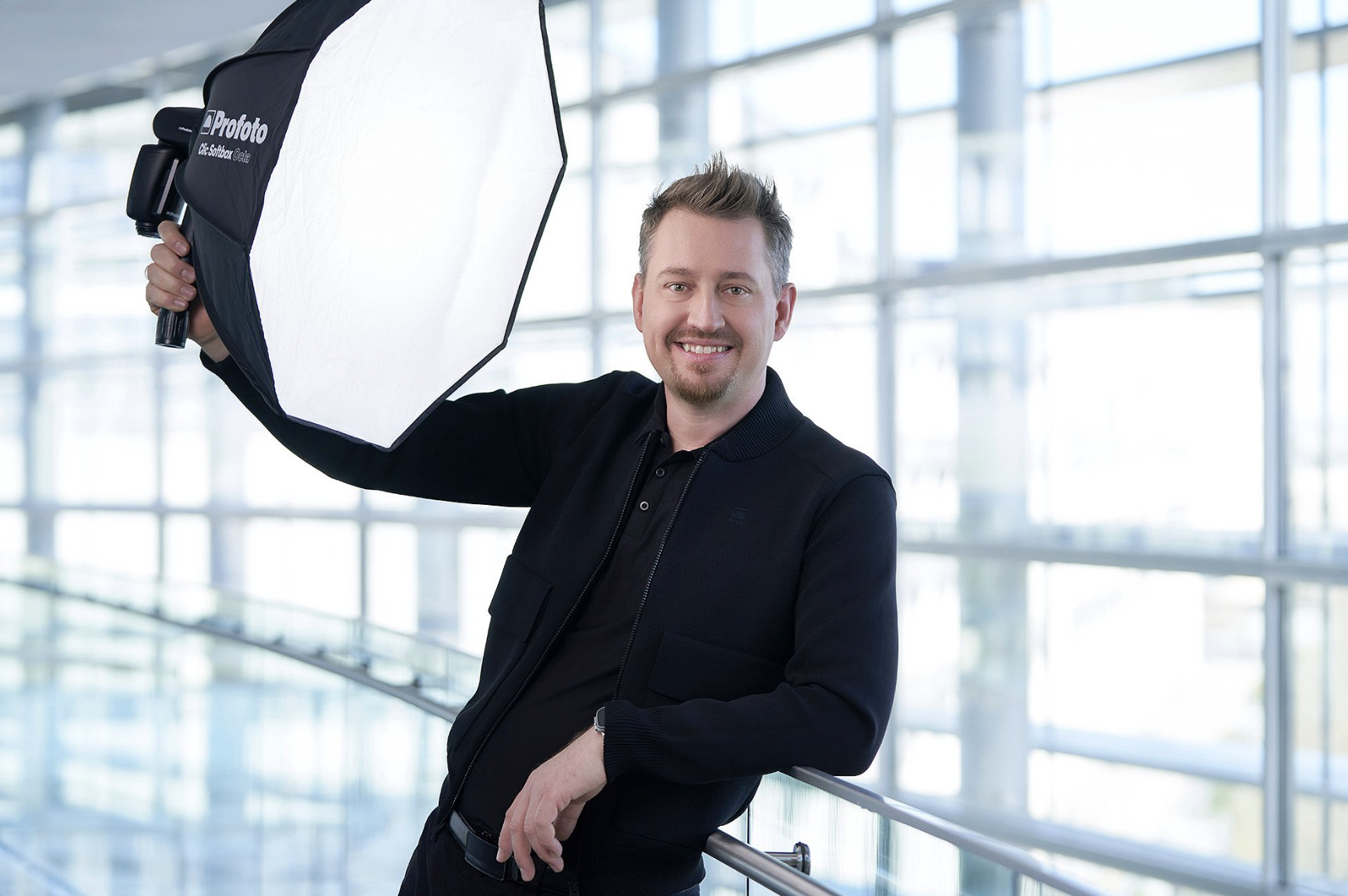 Ein lächelnder Mann posiert mit einem professionellen Fotoblitzlicht in einem hellen, modernen Gebäude mit großen Fenstern. Philipp Monihart Fotograf.