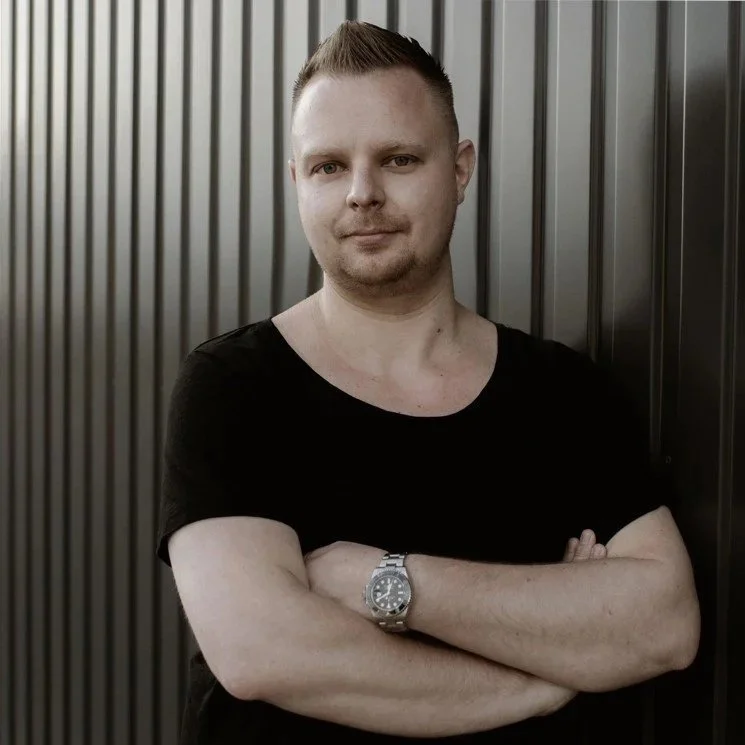 Mann mit Kurzhaarschnitt, schwarzem T-Shirt, Armbanduhr, steht mit verschränkten Armen vor einer dunklen Metallwand. Stefan Fried Geschäftsführer einer IT-Firma und Social-Media-Agentur.