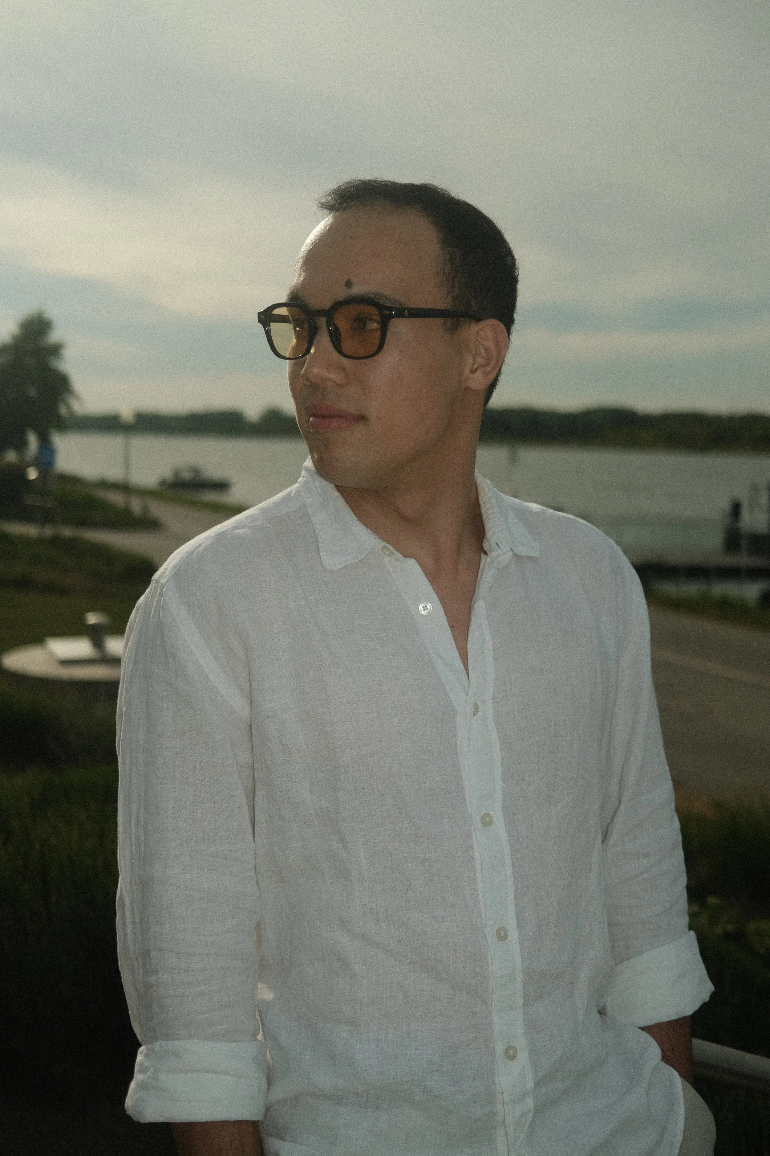 Ein Mann mit dunklen Haaren, Brille mit goldenen Gläsern, trägt ein weißes Hemd und steht an einem Fluss im Freien bei Sonnenuntergang.
