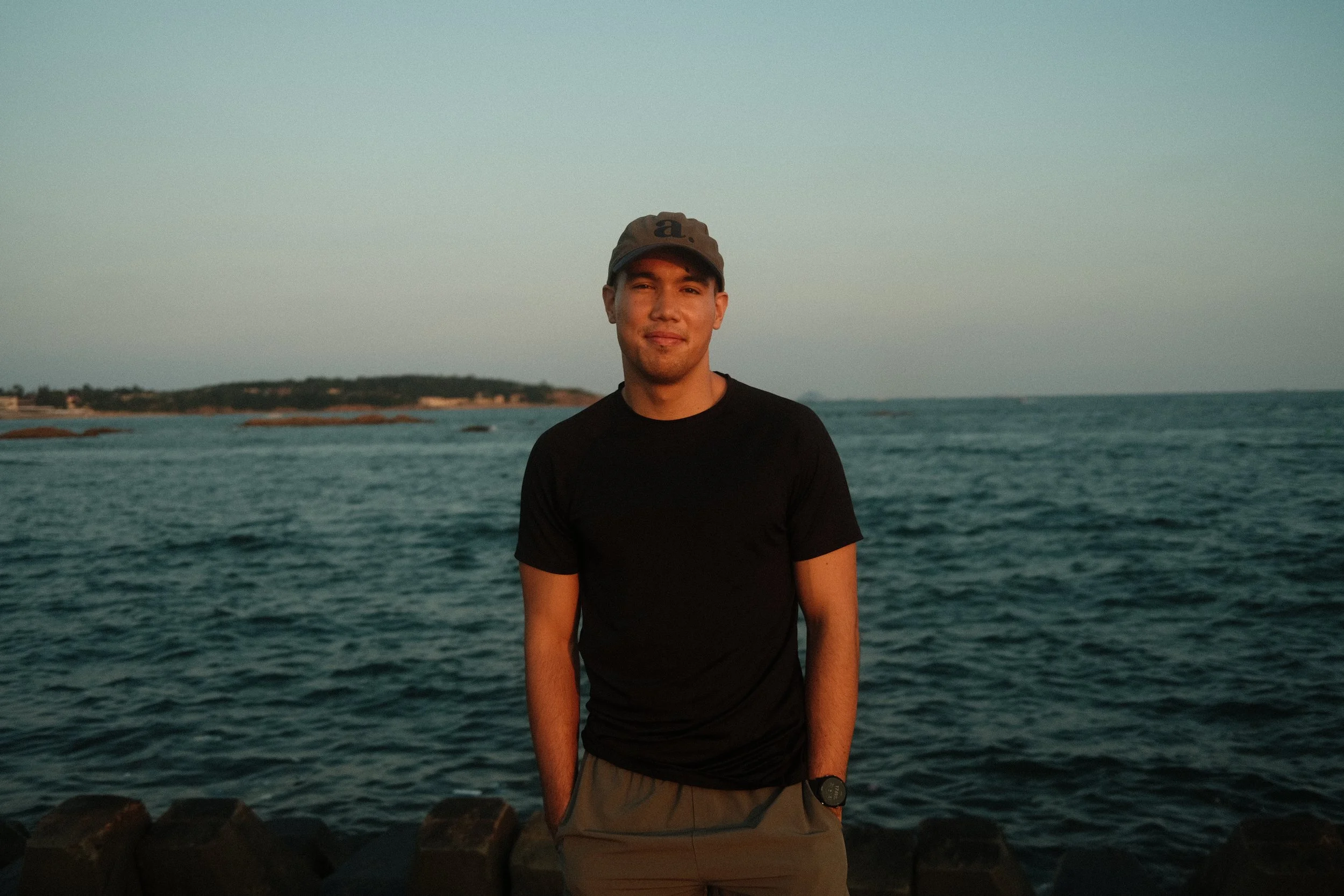 Junger Mann am Meer während des Sonnenuntergangs in China - Qingdao, trägt dunkles T-Shirt und eine Kappe.