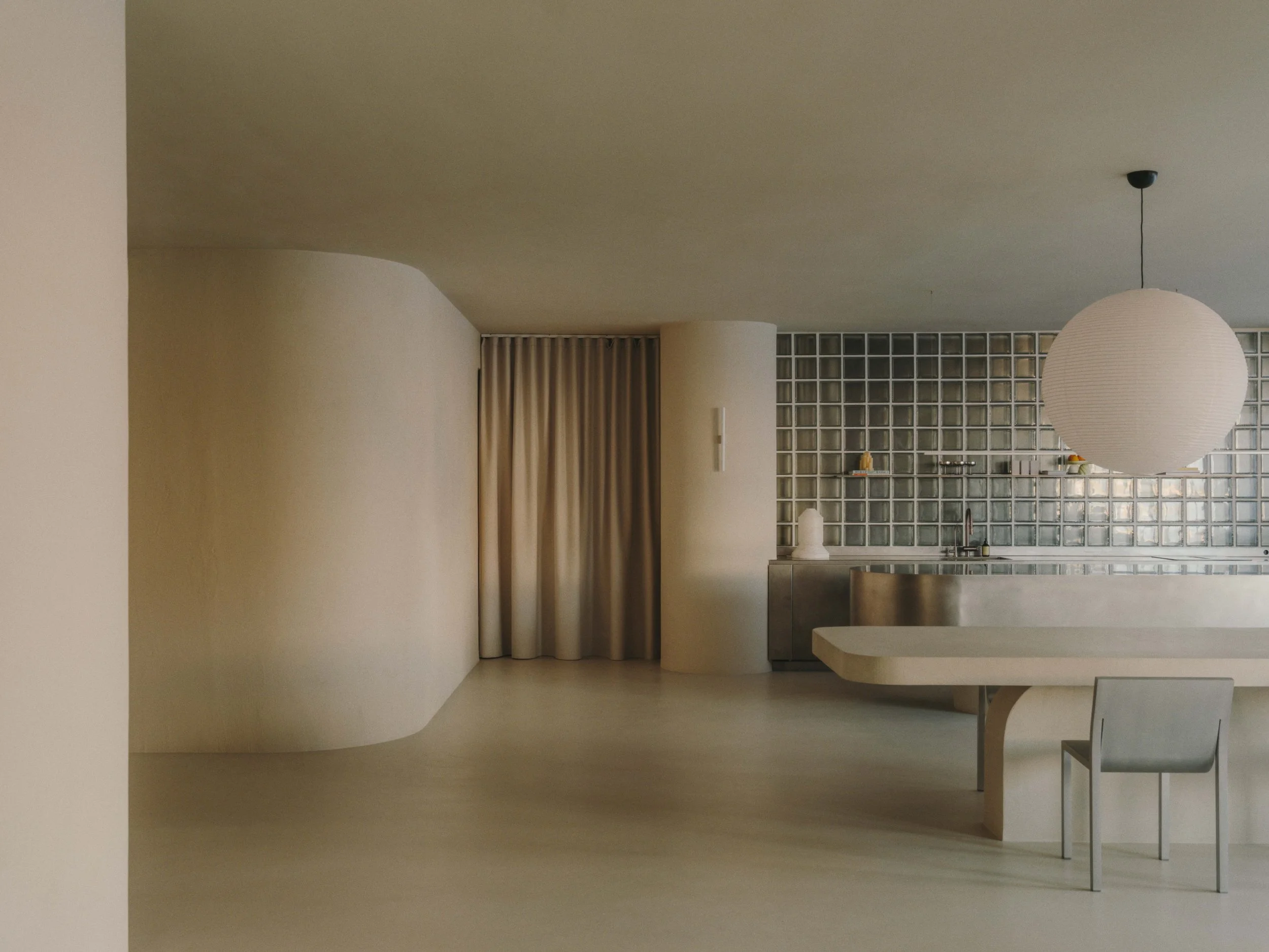 Minimalist kitchen with curved white walls, a round paper pendant light, a beige table, and a grid glass block backsplash.
