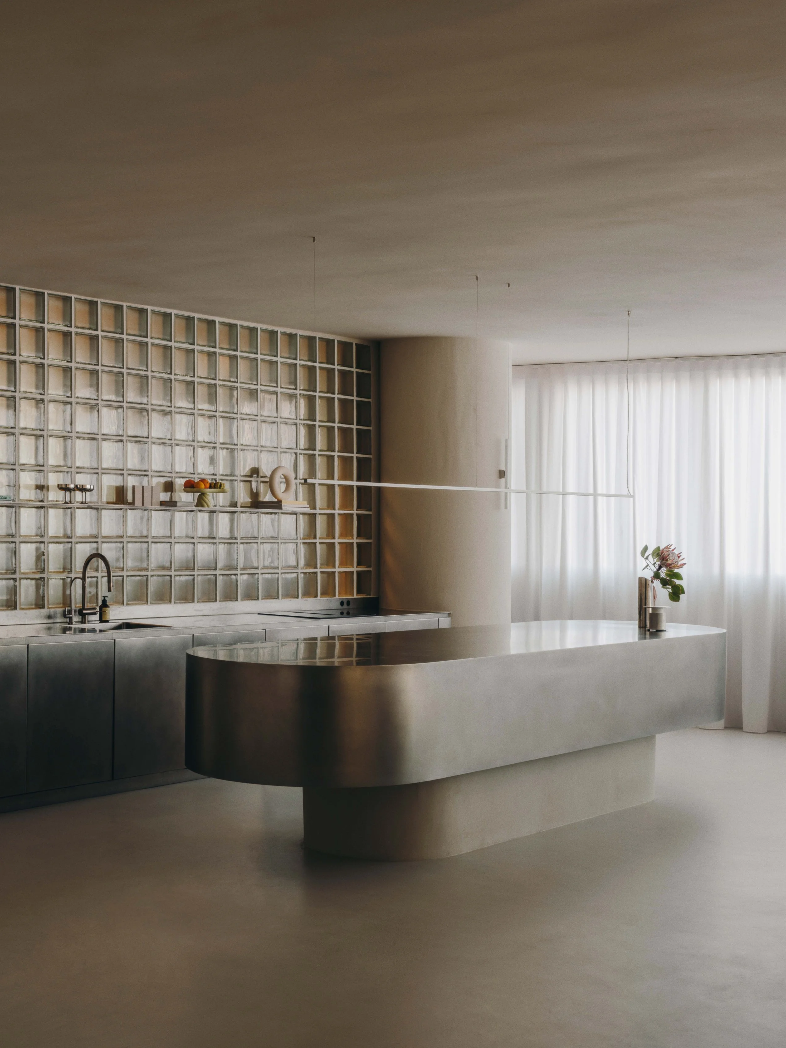 Minimalist modern kitchen with stainless steel counter and cabinetry, glass-block wall, and natural light from a large window with white curtains, decorated with a small plant.