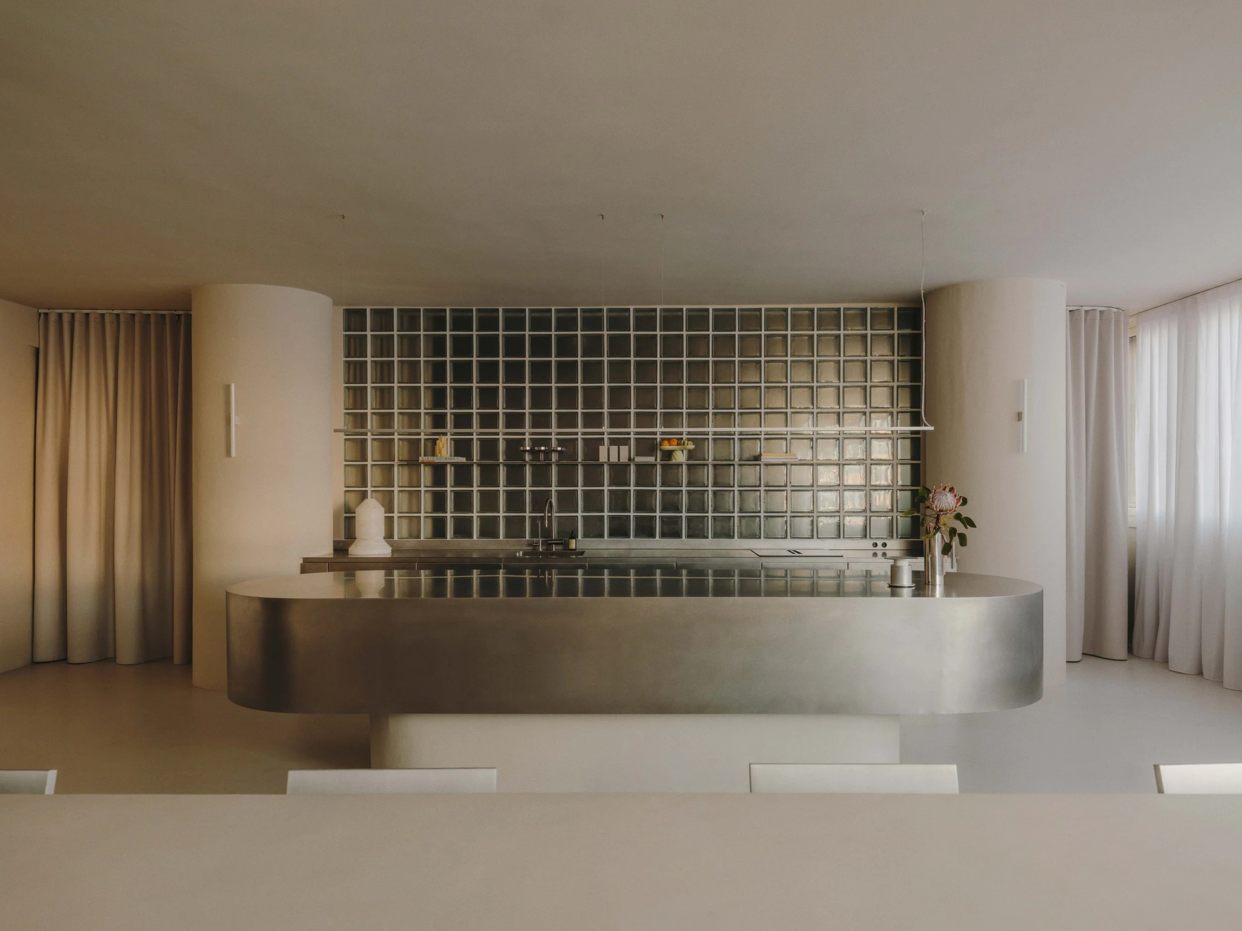 Minimalist kitchen with a curved metallic island, white walls, and a glass block backsplash. There are small items on shelves, and a flower in a vase on the counter, with curtains on both sides.