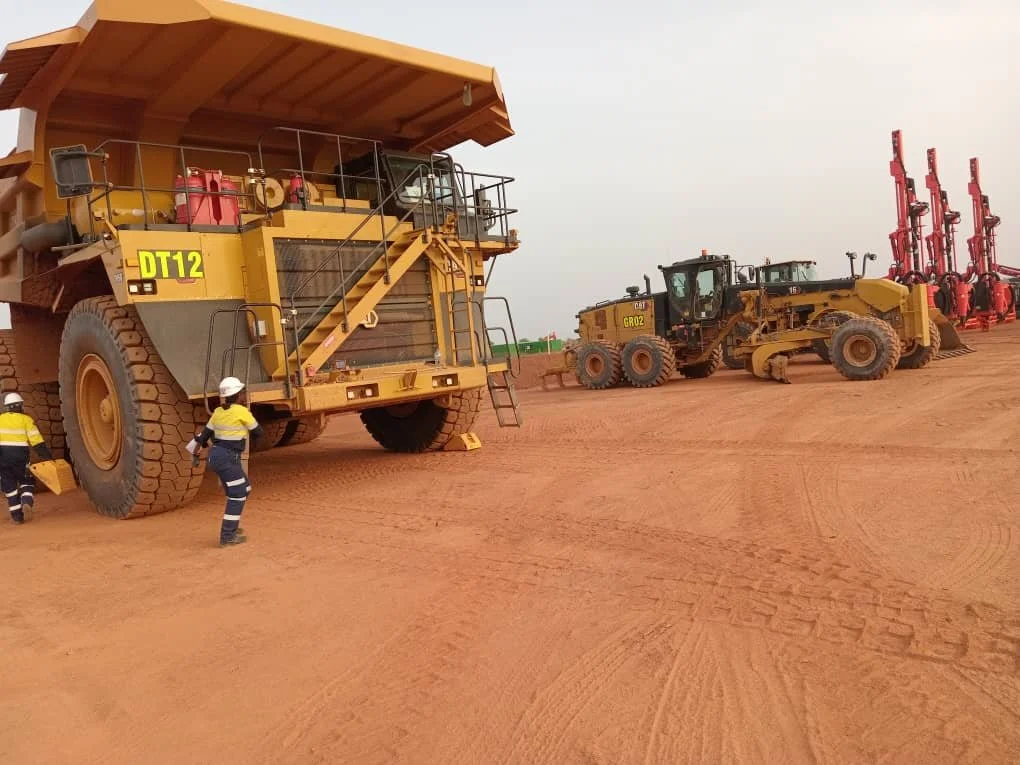 Workers inspecting large Caterpillar mining dump truck and smaller construction machinery on a dirt site with red soil.