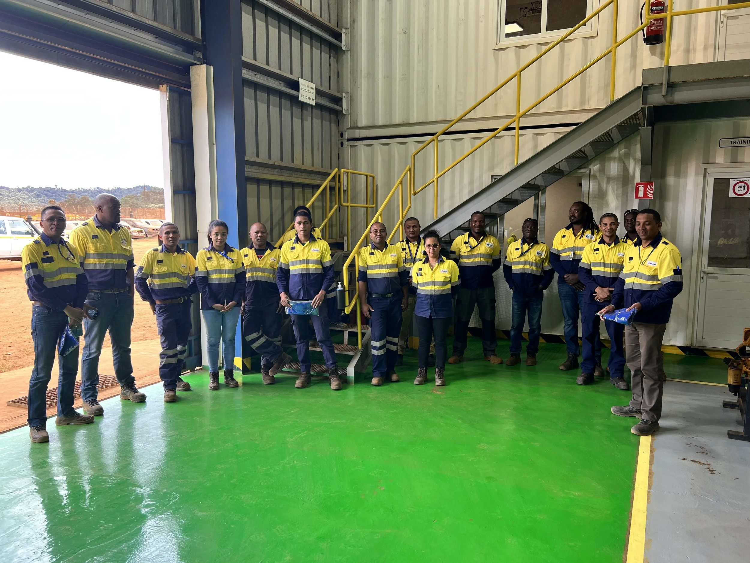 A group of people in yellow and navy safety uniforms standing inside a hangar or industrial building with a green floor, metal stairs, and large open garage doors revealing parked vehicles outside.