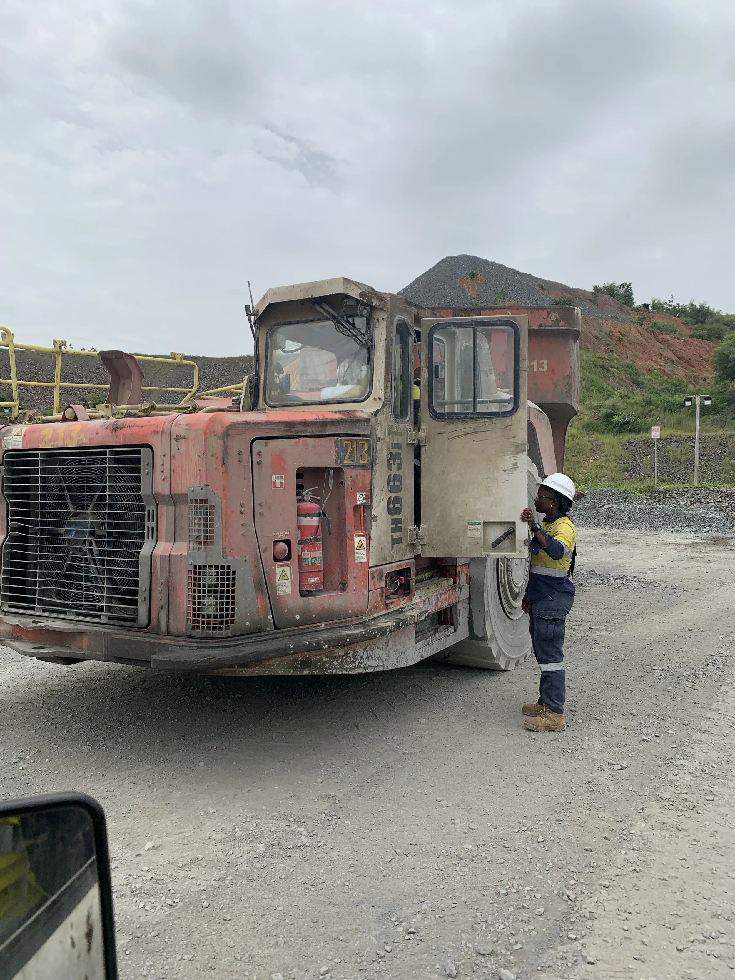 A construction worker wearing a white helmet and blue and yellow safety uniform inspecting a large red heavy-duty mining or construction vehicle at a mining site.