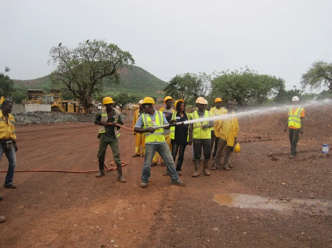 Group of construction workers and a woman with safety vests and helmets, testing a fire hose on a dirt construction site with machinery and trees in the background.