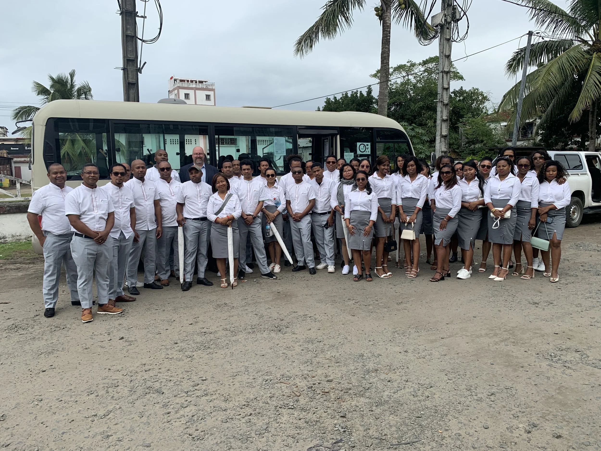 Group of men and women dressed in white shirts and gray skirts or pants standing in front of a bus and a white vehicle outdoors, with palm trees and electric poles in the background.