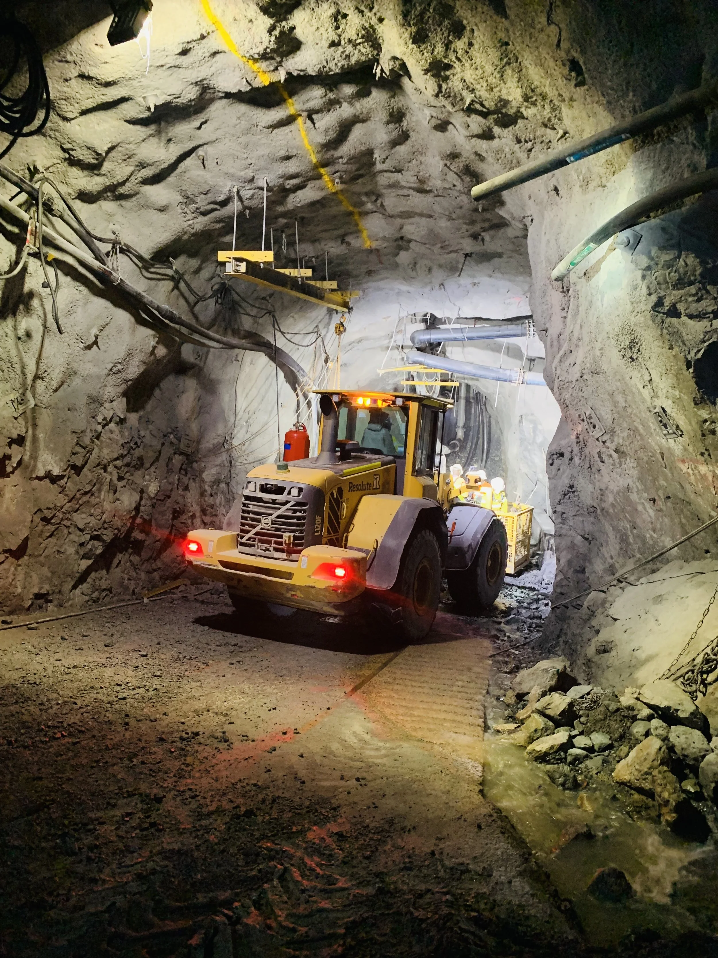 A yellow construction vehicle inside an underground mine tunnel, with rocky walls and pipes overhead, illuminated by lighting in the tunnel.