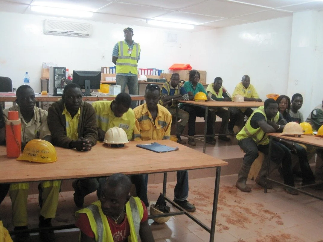 A classroom with multiple adult students wearing yellow safety vests, sitting at desks with safety helmets and water bottles, while an instructor stands at the front.