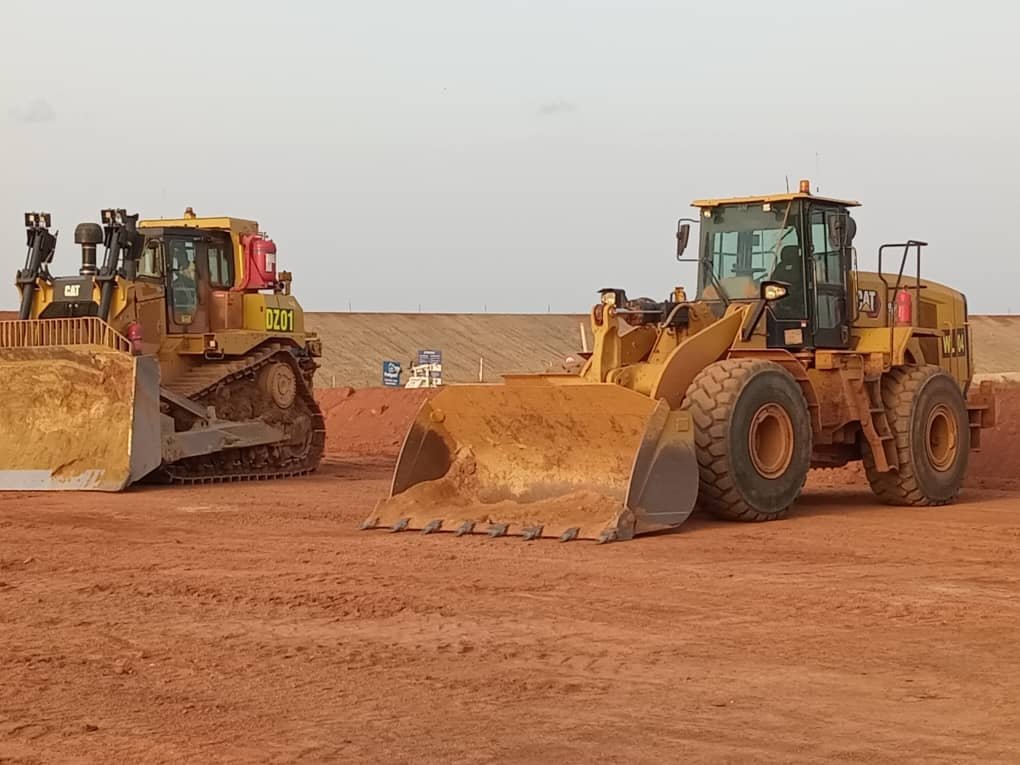 Two large bulldozers working on a construction site with red dirt, one in the foreground and one in the background under a cloudy sky.