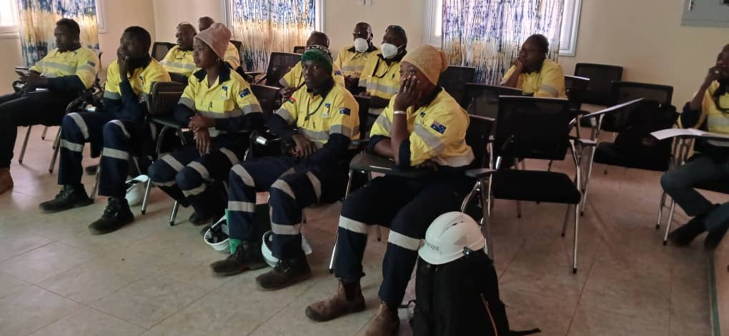 Group of workers in yellow and black uniforms sitting in a classroom or training room, some wearing masks and helmets, engaged in a training or meeting session.