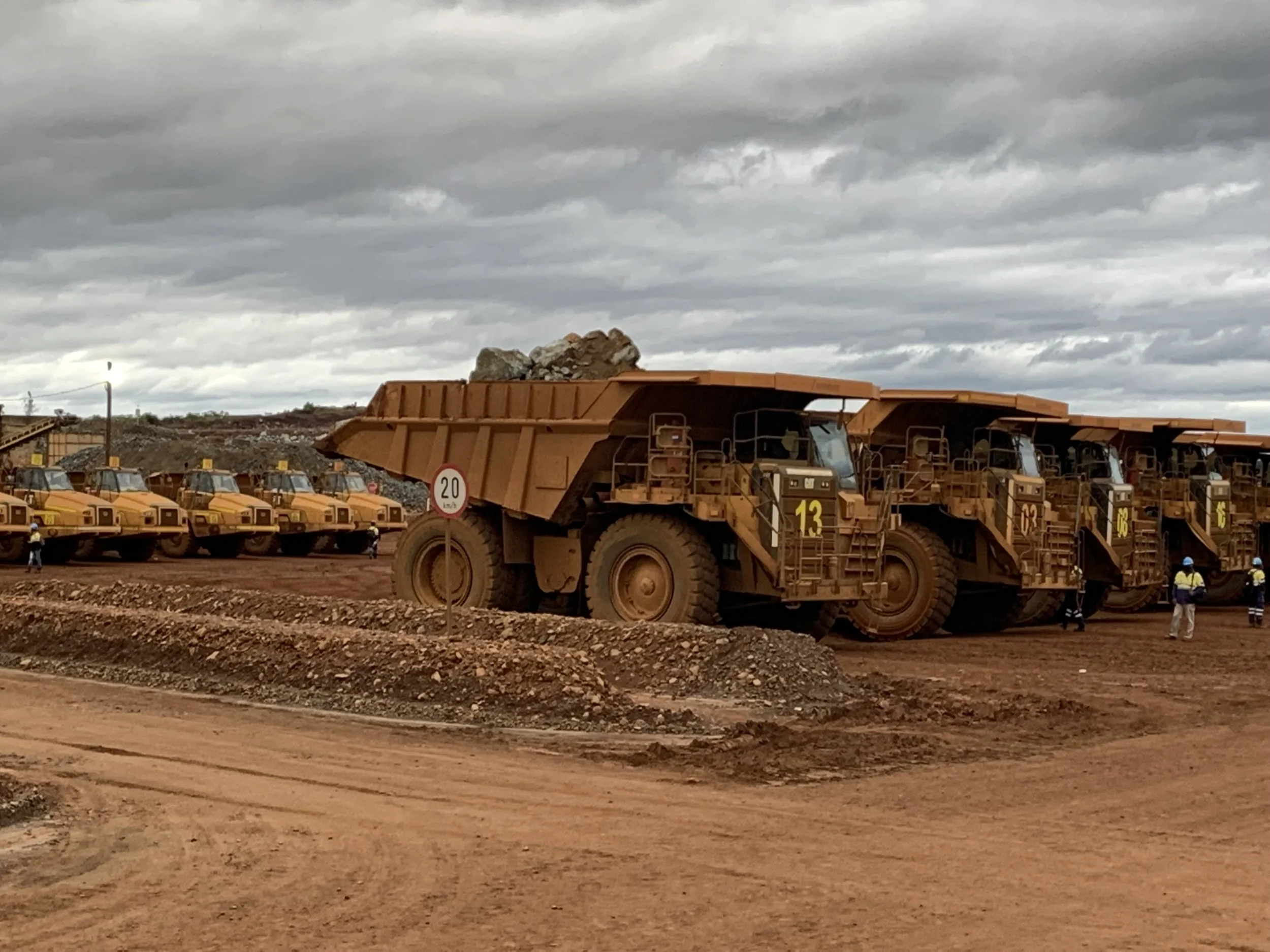 Large mining dump trucks parked in a row at a mining site on a cloudy day, with several workers in safety vests and helmets nearby.
