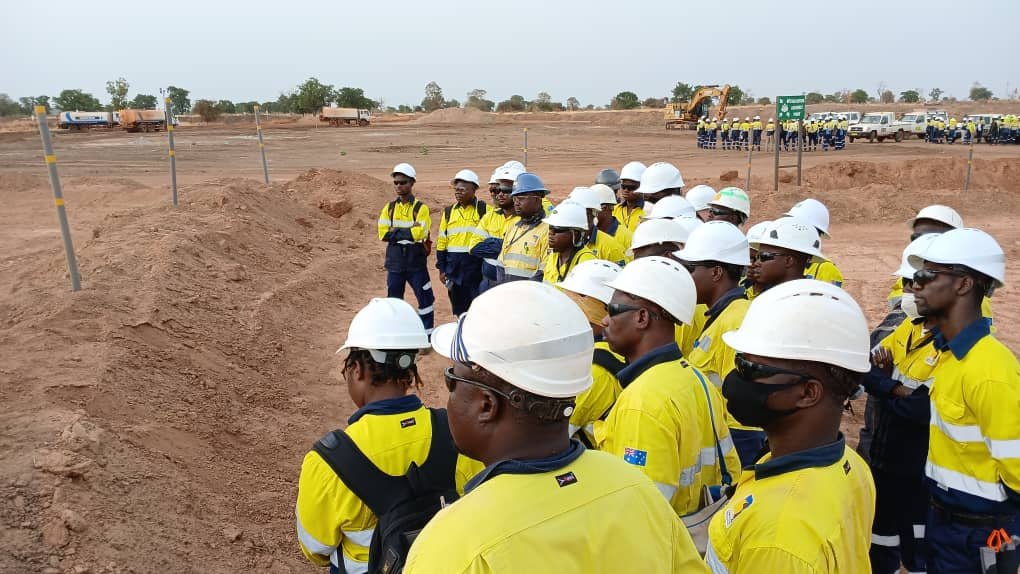 Group of construction workers wearing yellow shirts, white helmets, and safety gear standing in a line on a construction site with heavy machinery in the background.