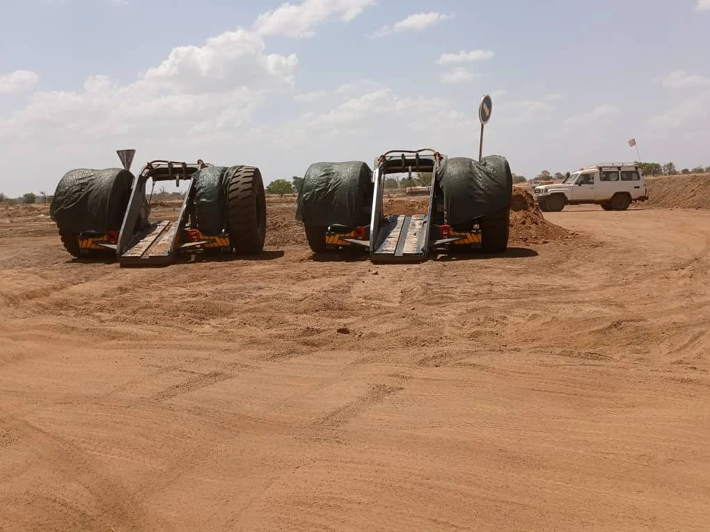 Two flatbed carts with large tires covered with tarps, parked on a dirt road with a white off-road vehicle in the background under a partly cloudy sky.