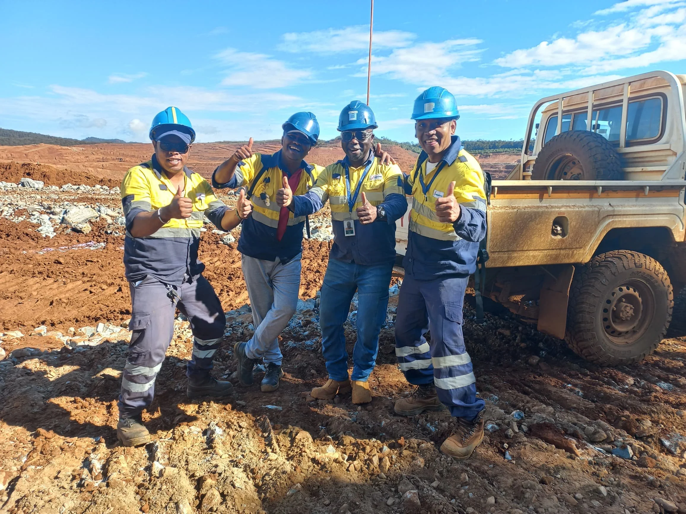 Four construction workers in yellow safety vests and blue helmets standing on dirt at a construction site, giving thumbs-up signs, with a beige vehicle in the background under a partly cloudy sky.