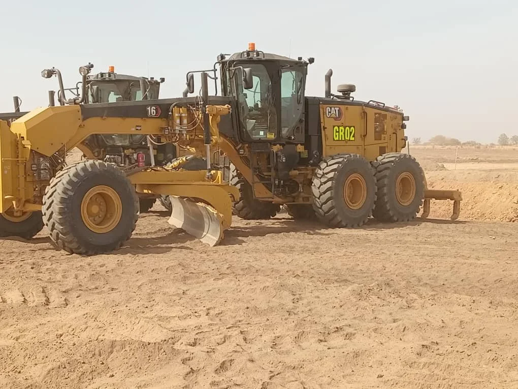 A yellow Caterpillar GR02 motor grader with large tires and a blade in the middle, situated on a dirt construction site under a clear sky.