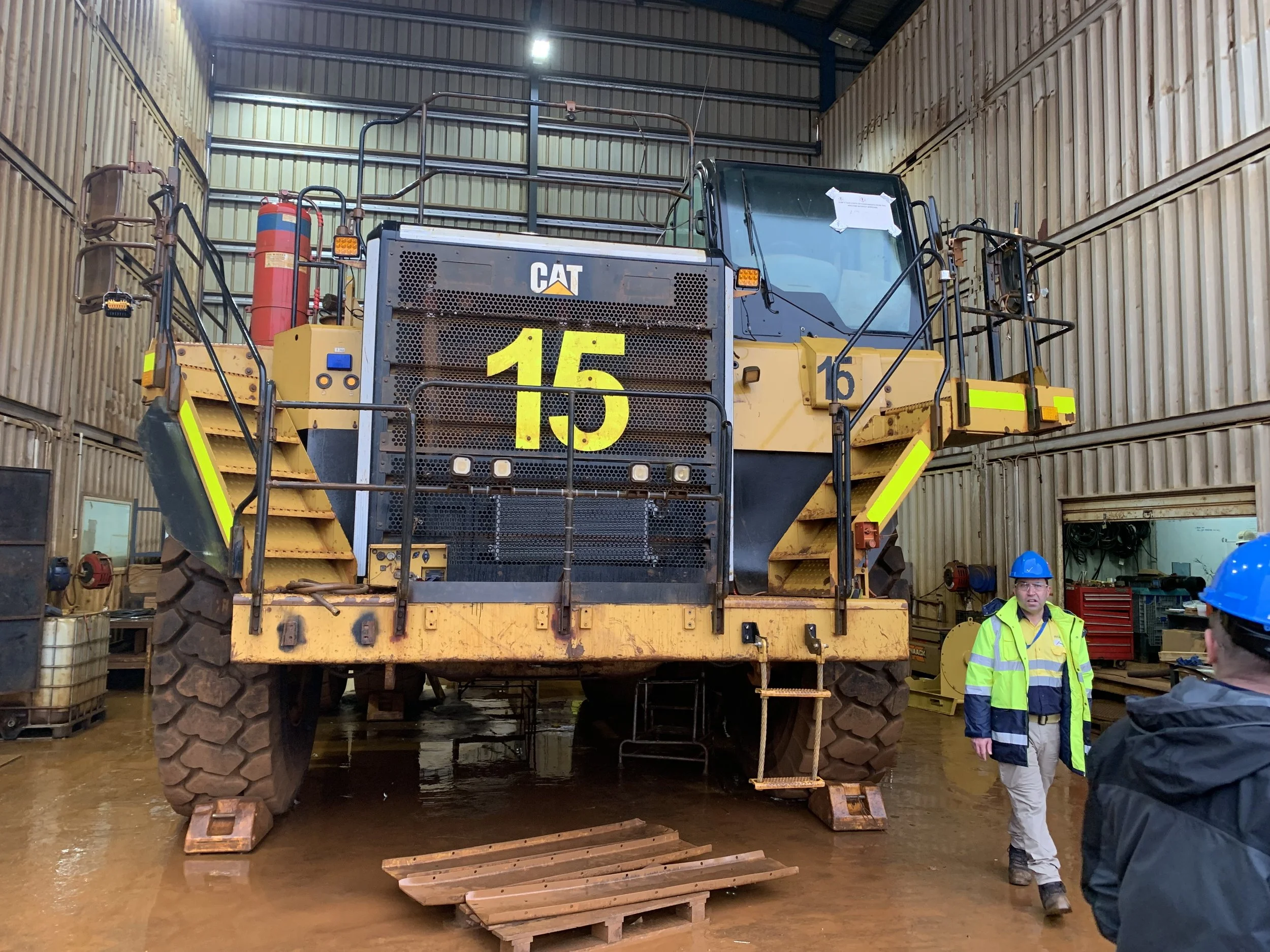 A large Caterpillar excavator inside a workshop, with two workers wearing blue helmets and high-visibility jackets inspecting it.