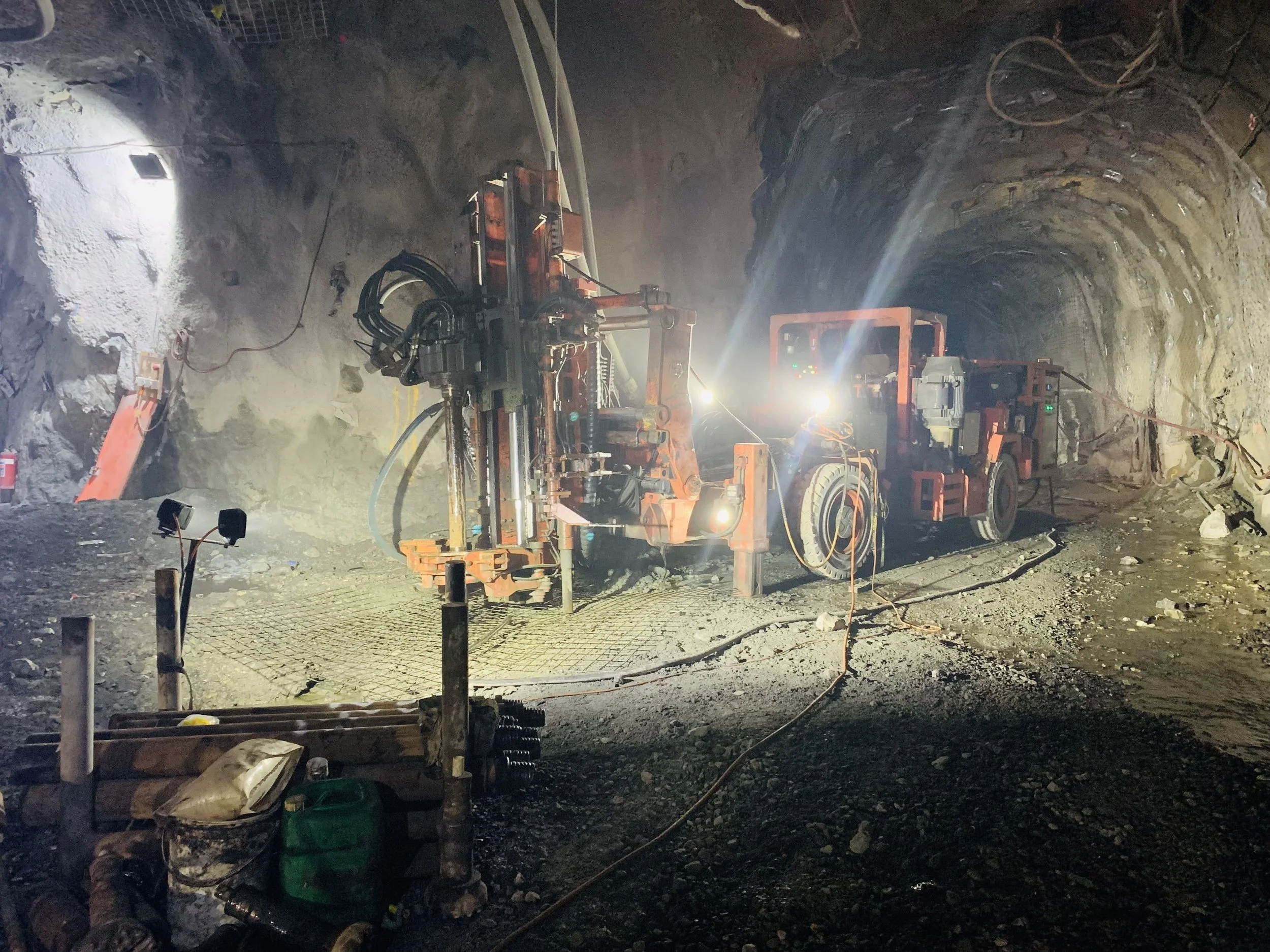 Underground mining tunnel with large drilling machines and equipment, illuminated by bright lights, dust and debris on the ground.