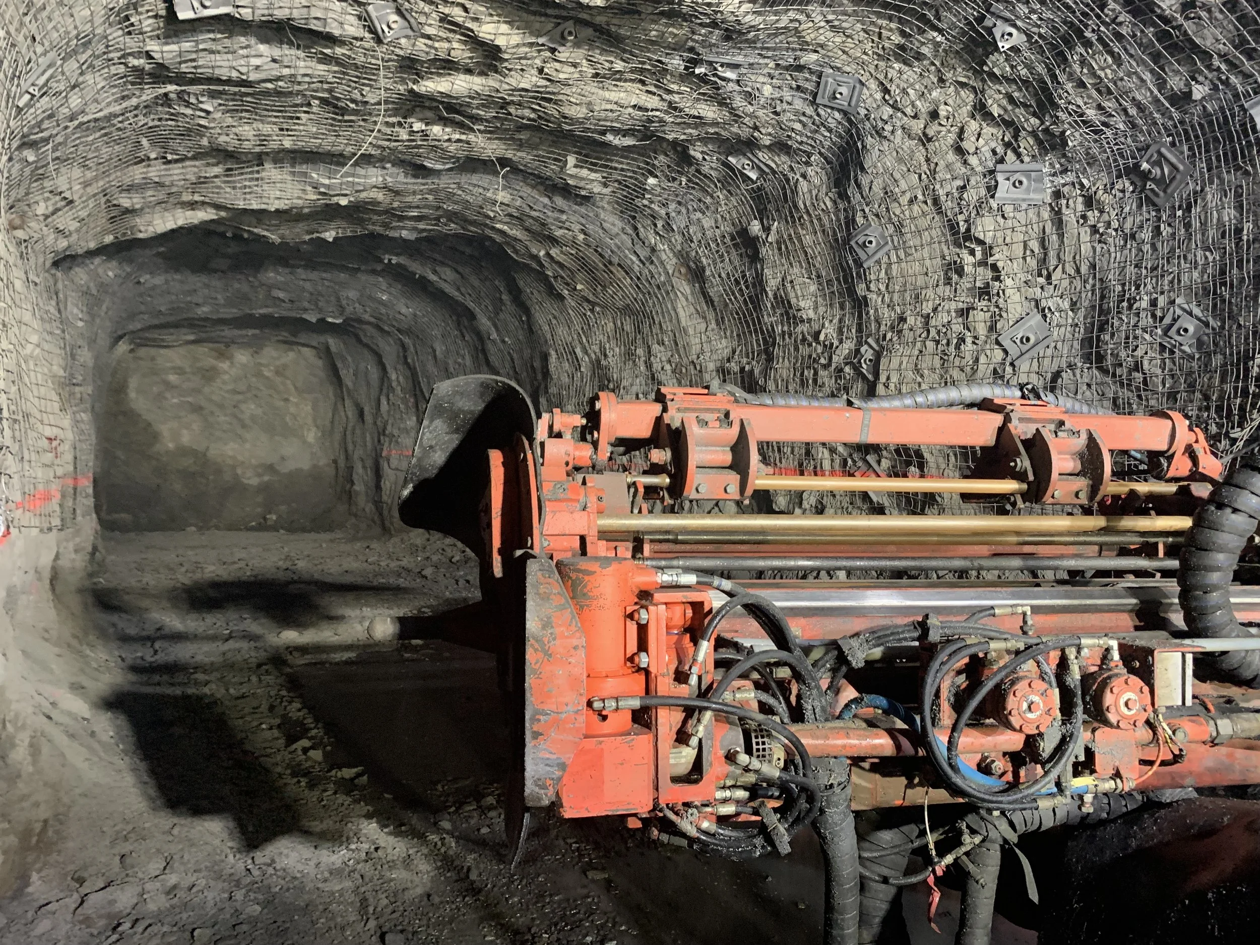 Interior view of a tunnel under construction, showing a tunnel boring machine with a large cutting head and machinery on a dirt floor, with rock walls and netting on the tunnel ceiling.