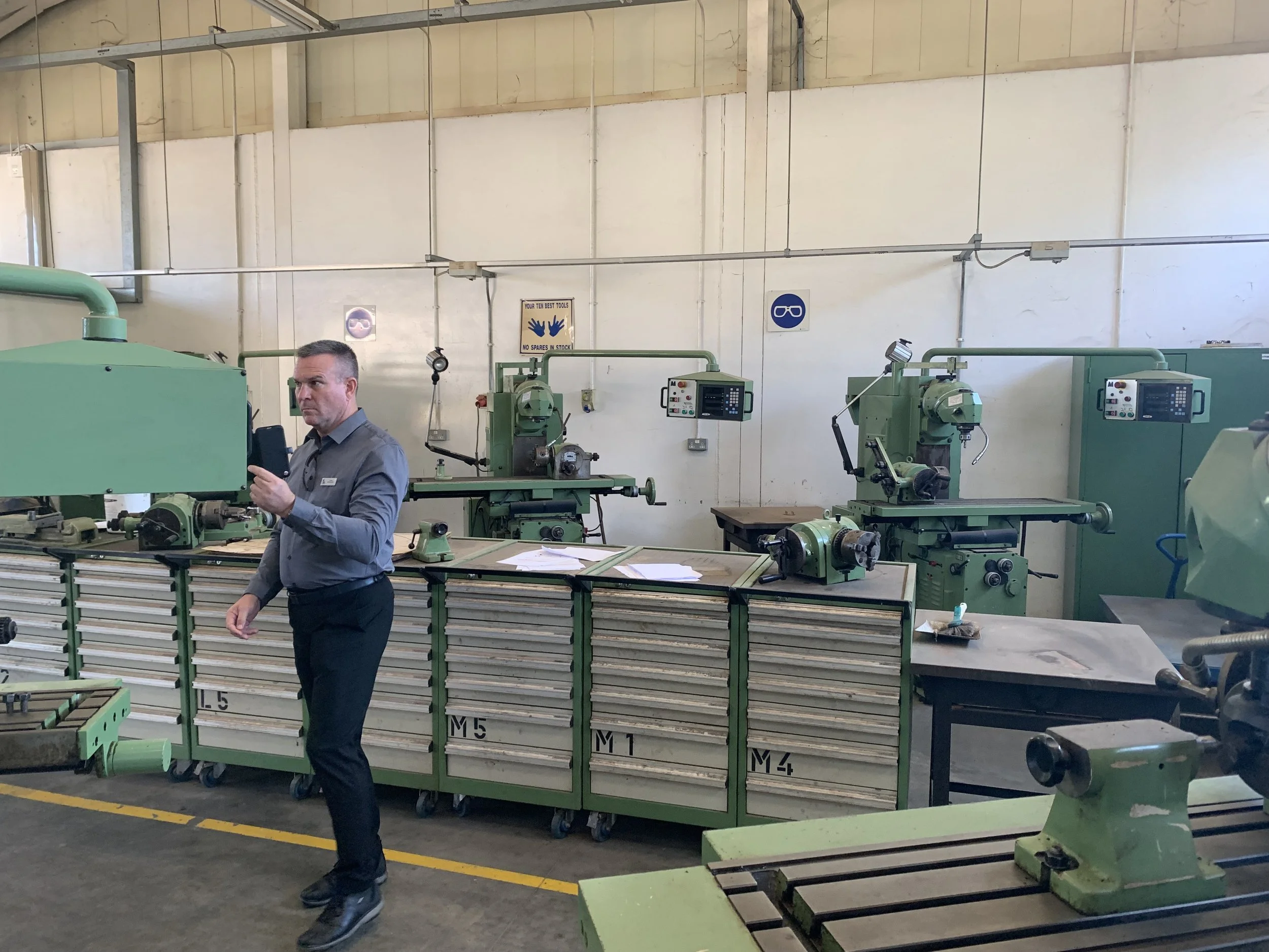 A man in a gray shirt and black pants standing in a machine shop, holding his phone. The shop has several green milling machines and metal tool drawers, with safety signs on the wall.