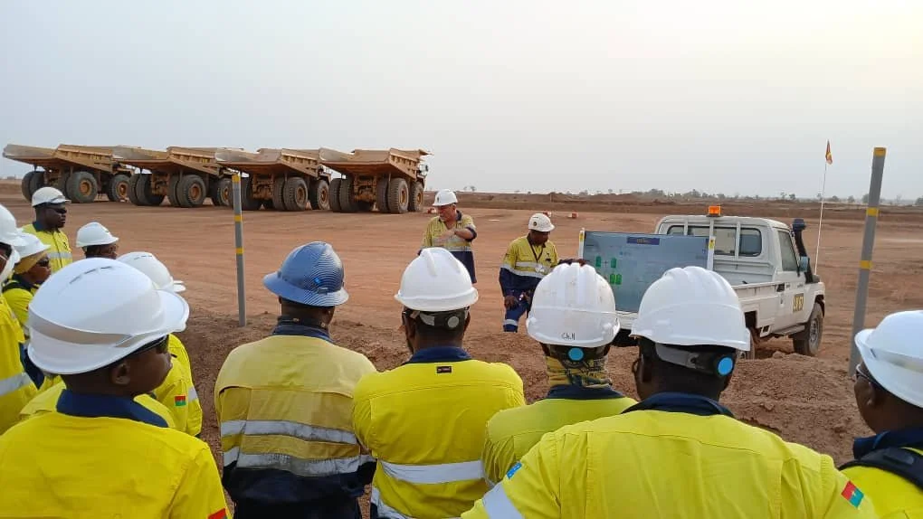 Construction workers in yellow uniforms and white safety helmets gathered on a dirt construction site, with a white pickup truck and large heavy machinery in the background.