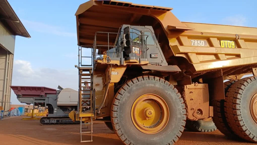 Close-up of a large yellow Caterpillar dump truck parked outside, with a smaller machine attached to it, under a partly cloudy sky.