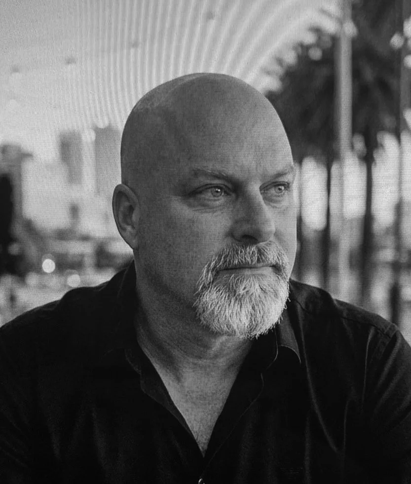 A black and white photo of a bald man with a beard, wearing a dark shirt, sitting outdoors near palm trees with blurred background.