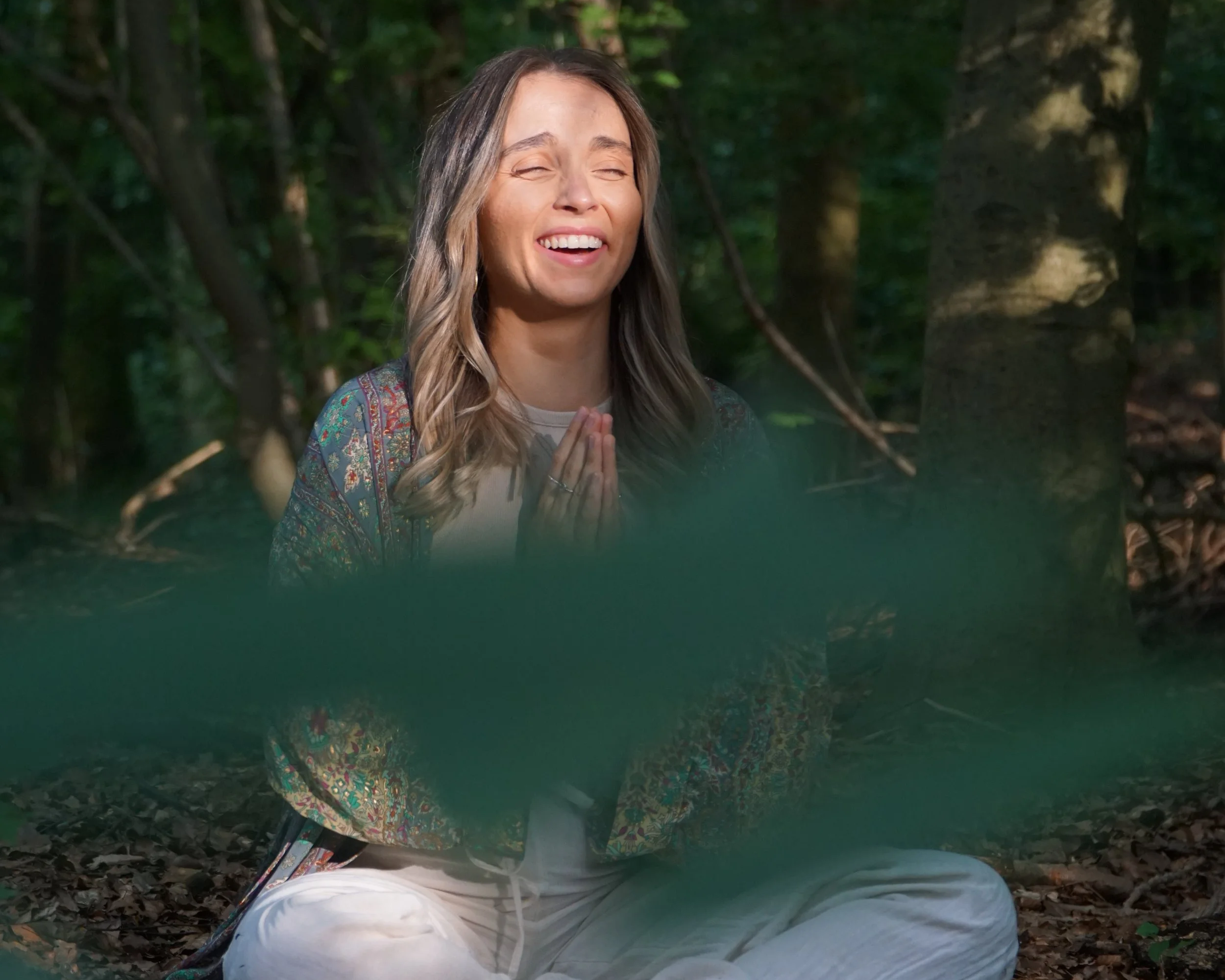 A woman sitting cross-legged in a forest, with her hands in prayer position, smiling with eyes closed.