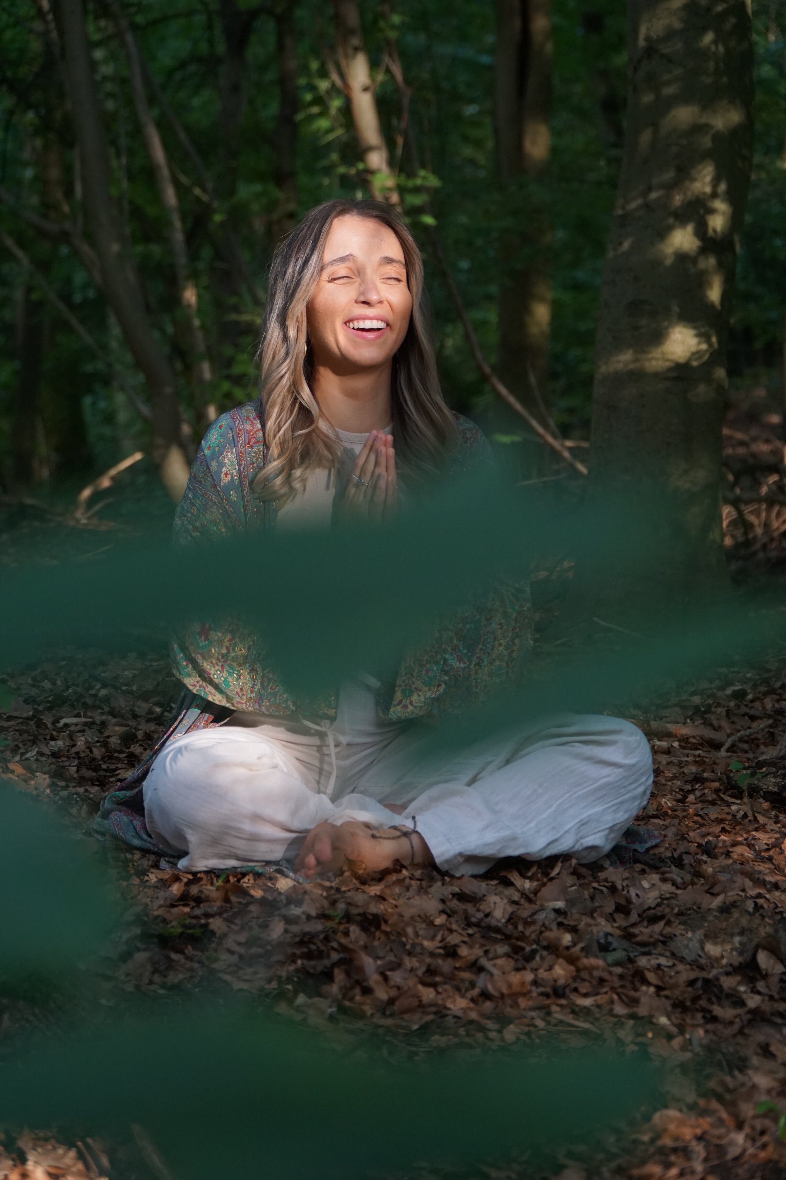 A woman with long wavy hair, sitting cross-legged on the forest floor with her hands in a prayer position, smiling with eyes closed, surrounded by trees and leaves.