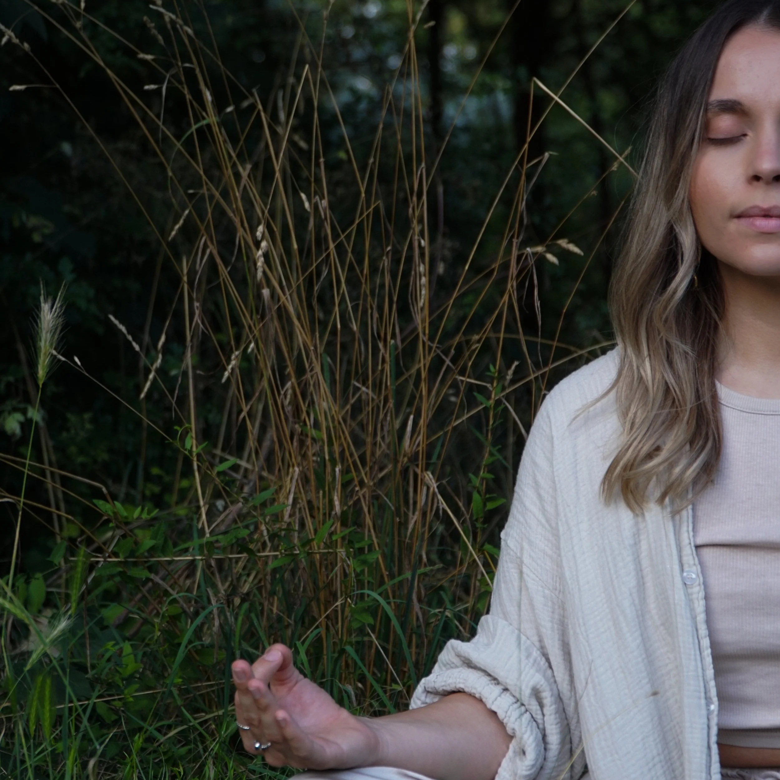 A woman meditating outdoors among tall grass and greenery, with her eyes closed and hand resting on her knee.