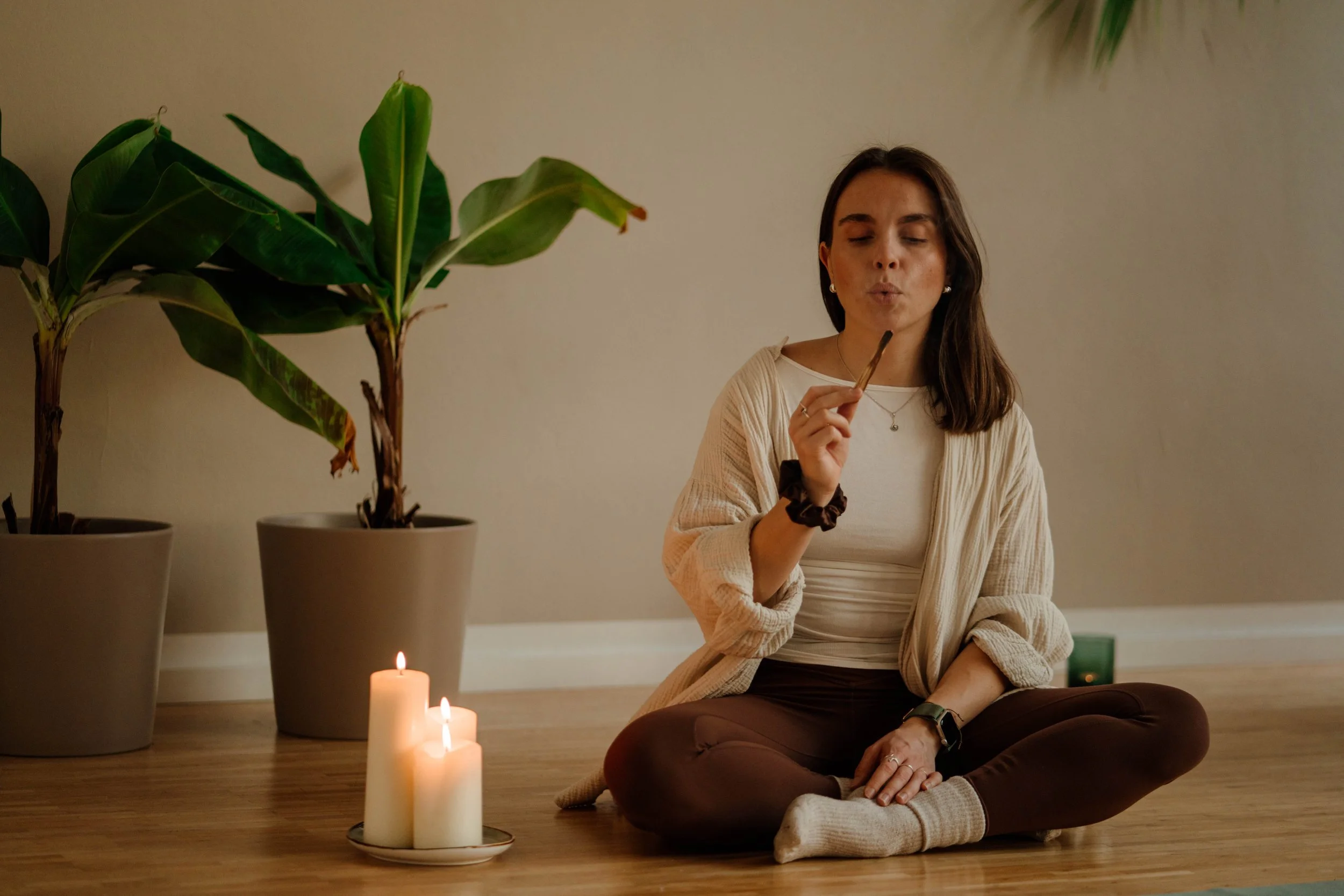 A woman sitting cross-legged on a wooden floor, holding a lit incense stick, with candles burning nearby and large potted plants in the background.