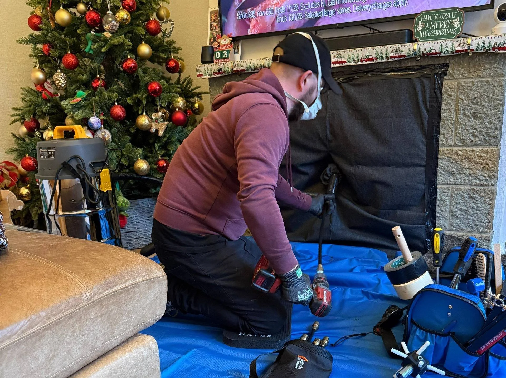 A man wearing a cap, glasses, face mask, hoodie, and gloves kneeling on a blue mat, using power tools to work on a fireplace or chimney in a living room decorated for Christmas with a decorated Christmas tree, and tools and equipment nearby.