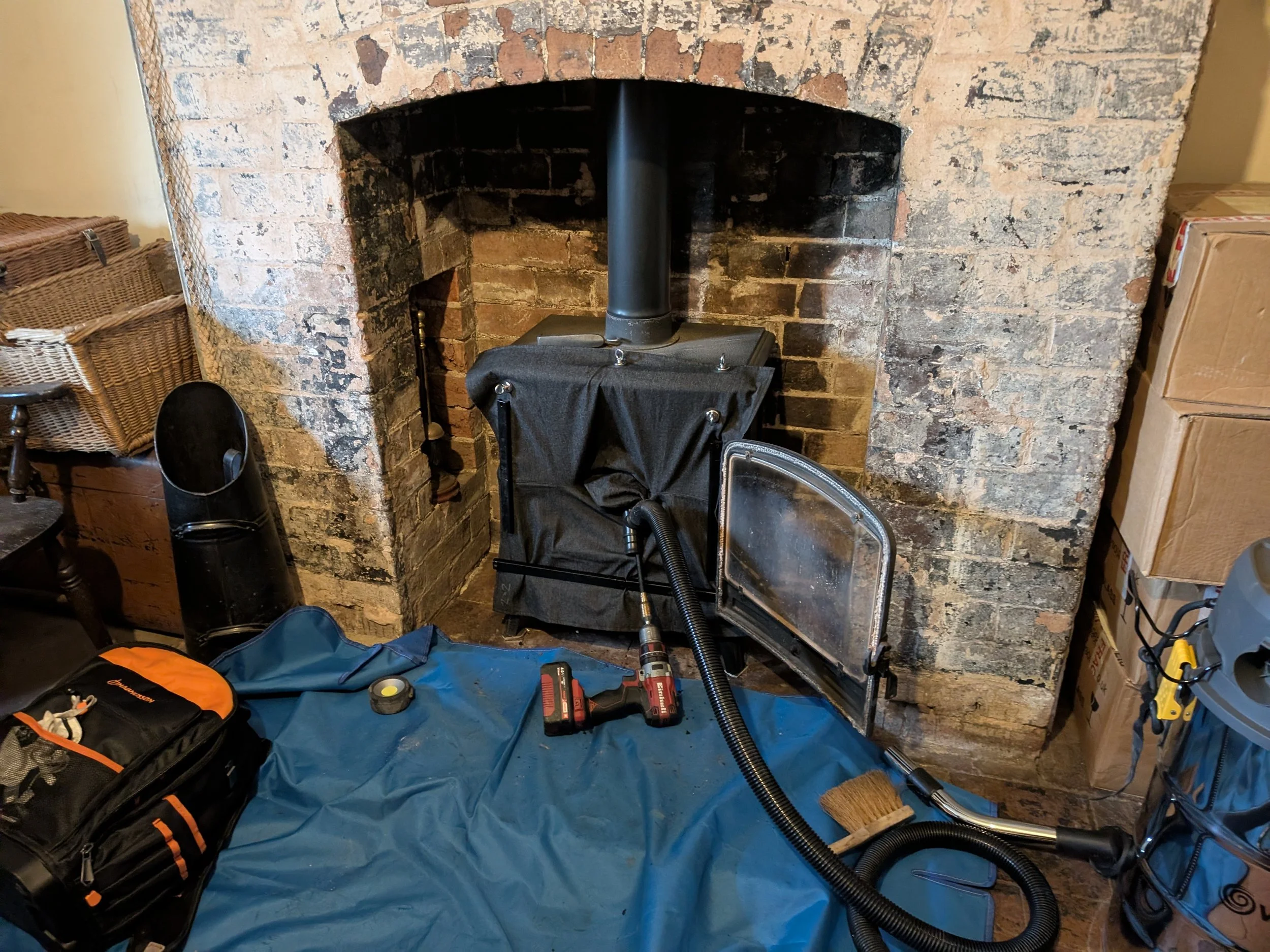 Inside a room with a brick fireplace and a wood stove being installed. Tools, a drill, a broom, and a blue tarp are on the floor. Cardboard boxes are stacked on the right side.