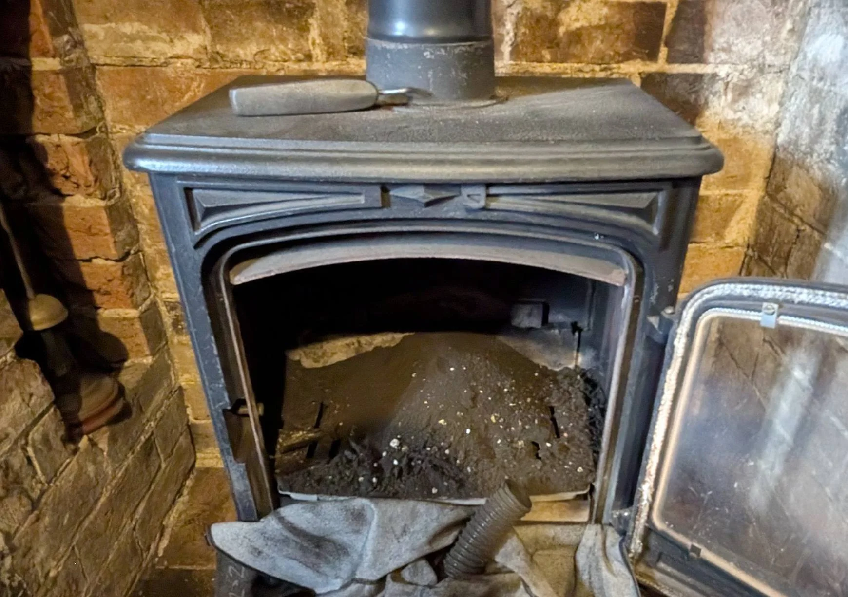 A black cast iron wood stove with a glass door, filled with ashes and soot, surrounded by brick walls.