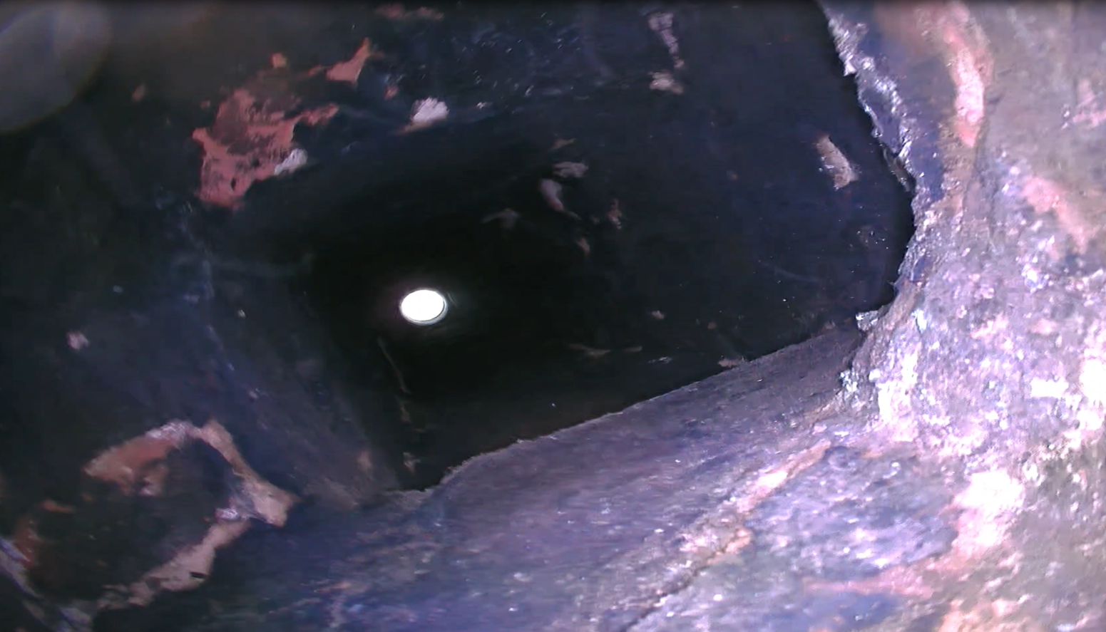 Looking down into a dark, deep, cylindrical hole in the ground with rough rocky surfaces, with a small bright light at the bottom.