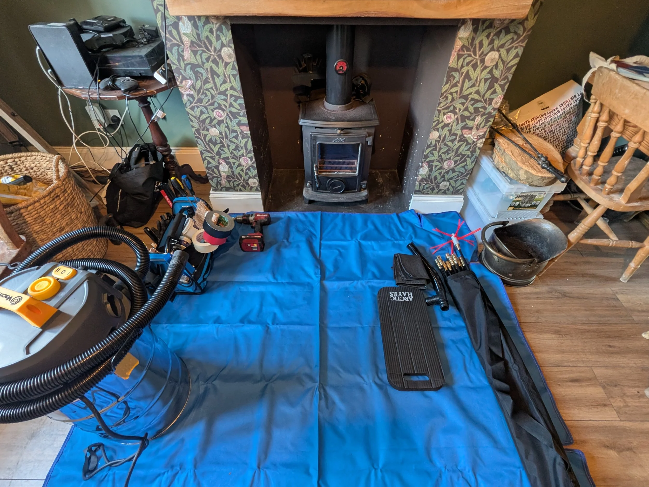 This room appears to be a workshop or repair area with various tools and equipment. There is a vacuum cleaner on a blue mat, a set of screwdrivers, a black carrying case, and various other wiring and tools. The background features a fireplace with a 