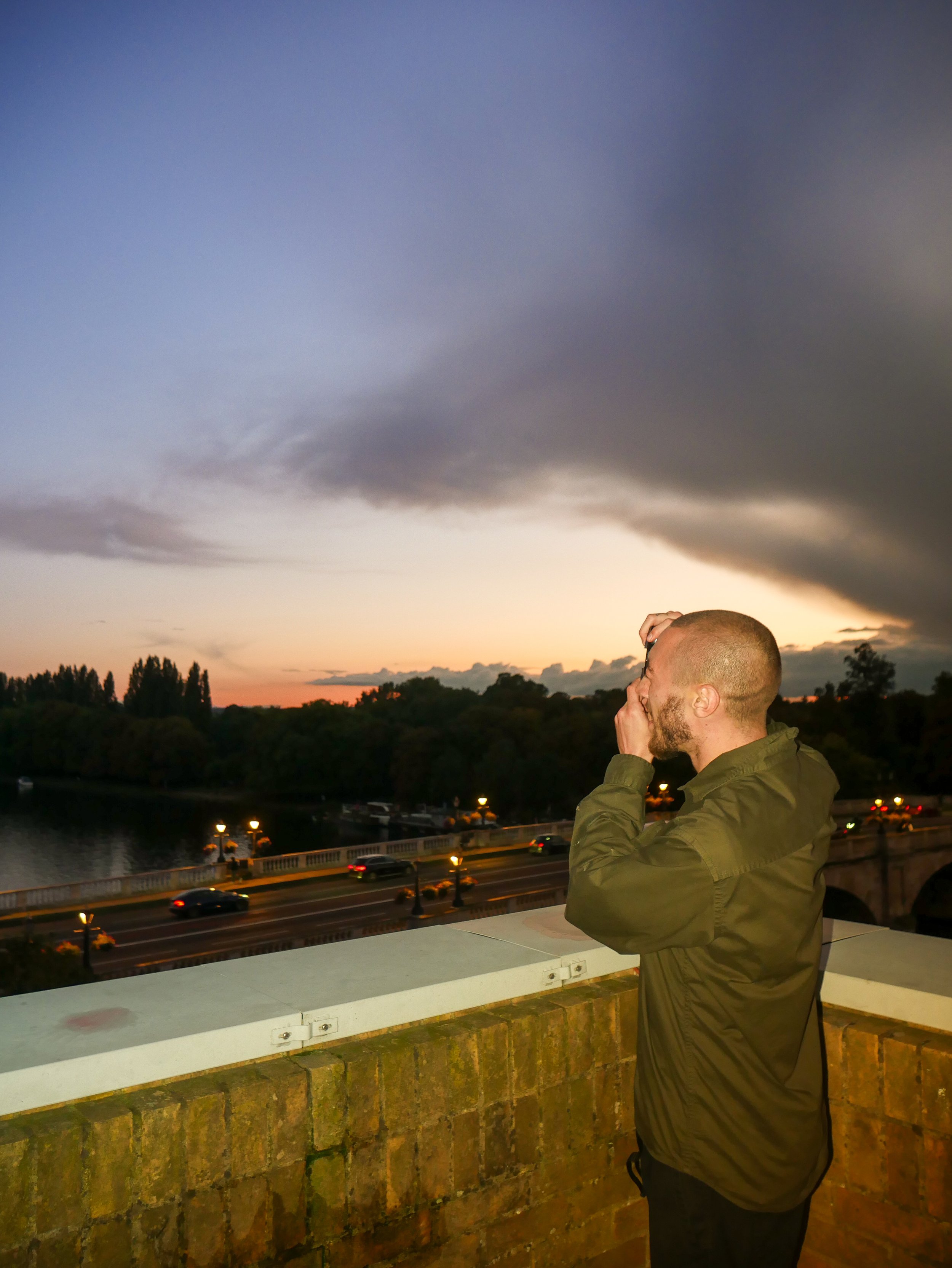 Man photographing city skyline at sunset from a bridge, capturing urban atmosphere and light