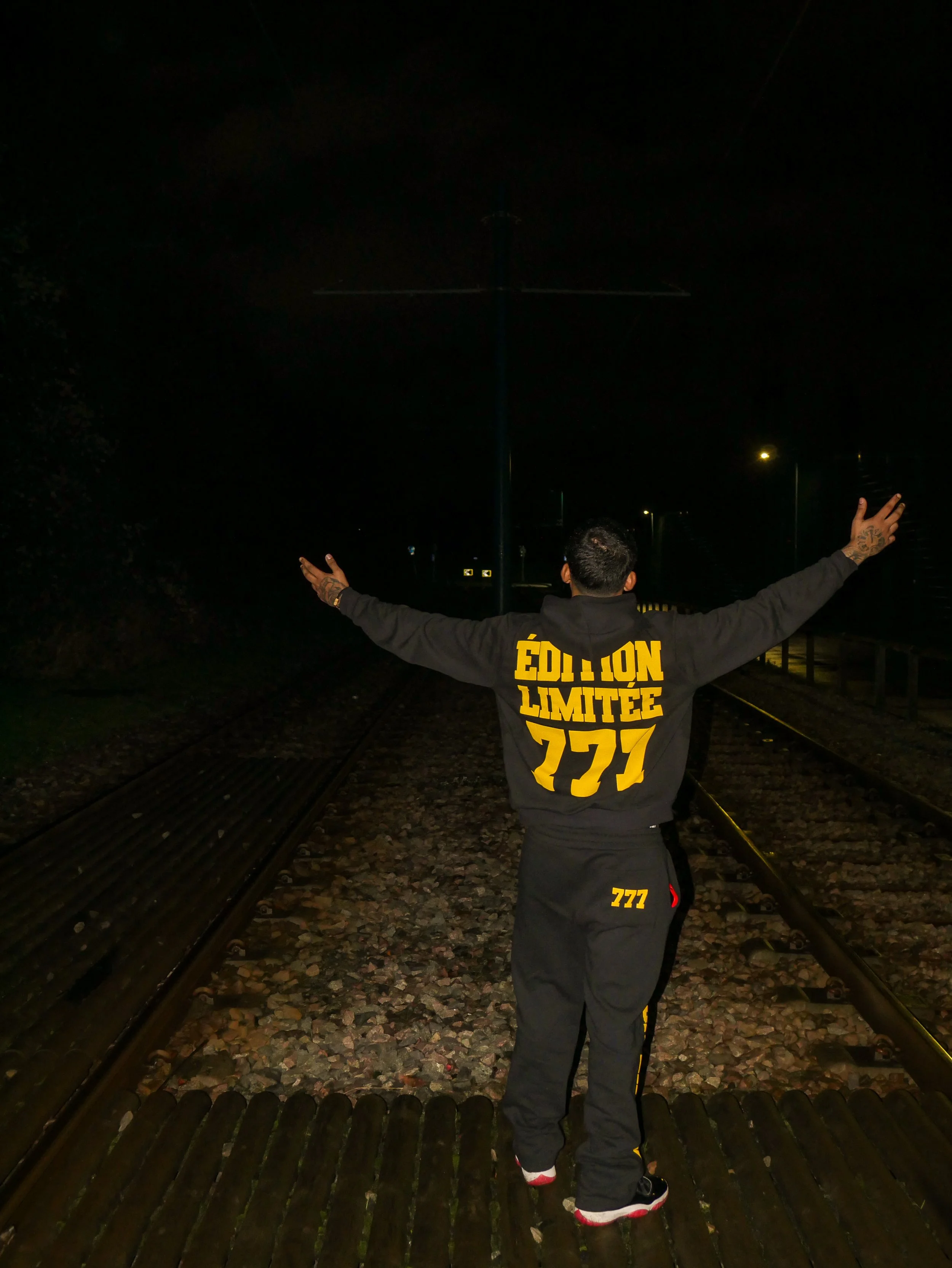 Man standing on railway tracks at night with arms outstretched, wearing a black tracksuit with yellow “Édition Limitée 777” text on the back.