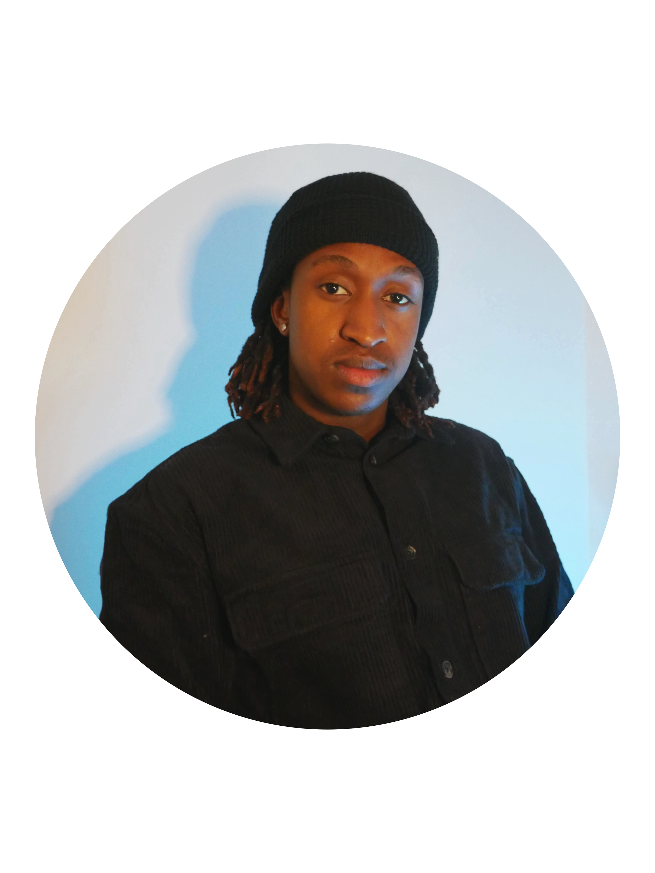A young man with dreadlocks wearing a black beanie and black button-up shirt, standing in front of a light-colored wall with blue lighting.