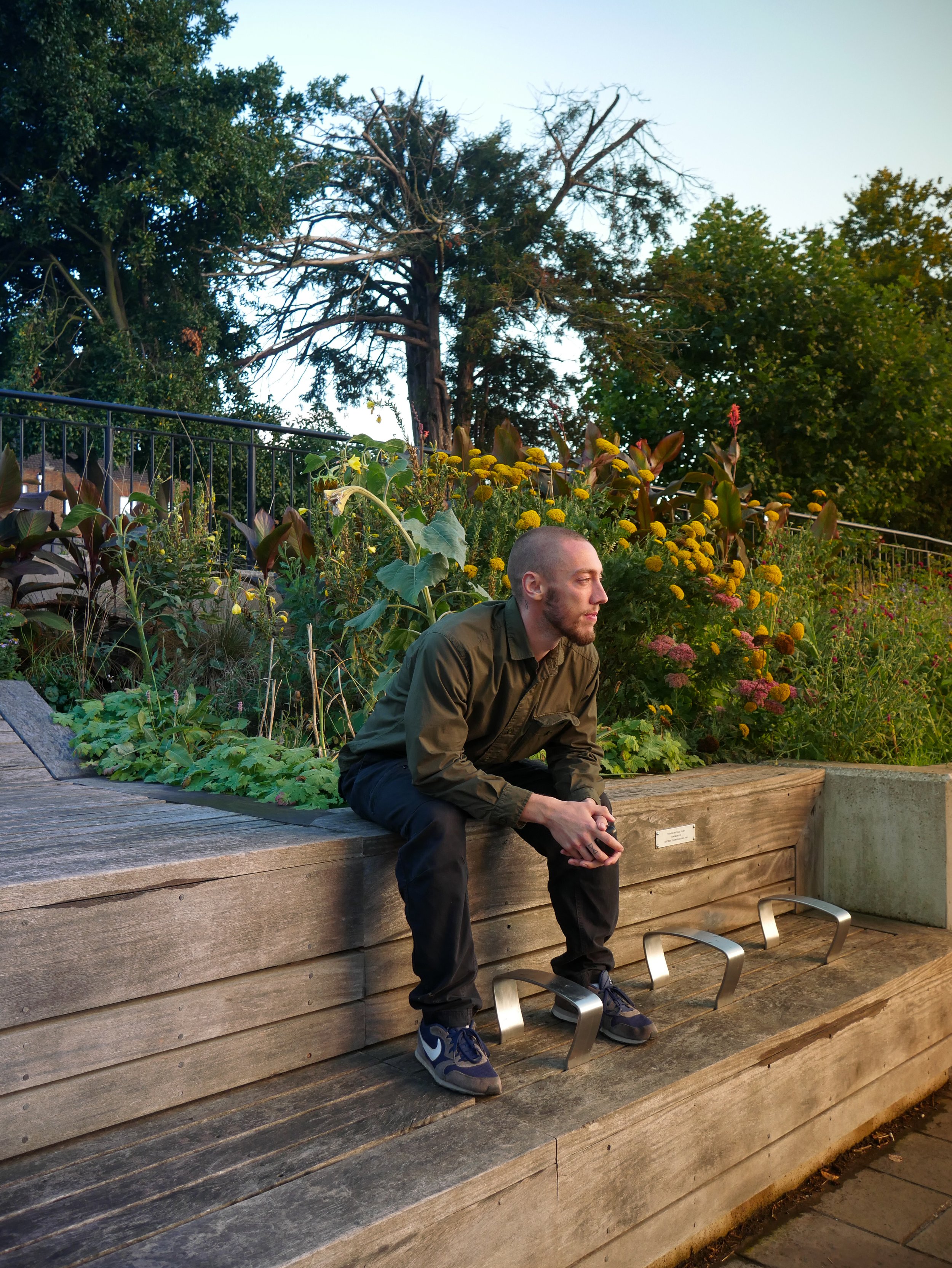 A man with a shaved head and beard sitting on a wooden bench in a park, surrounded by flowers and greenery, during sunset.