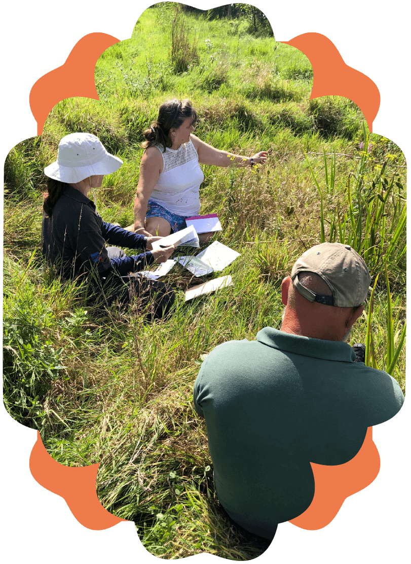 A woman and a girl sitting in tall grass, examining plants and taking notes, while a man with a cap sits nearby, observing.