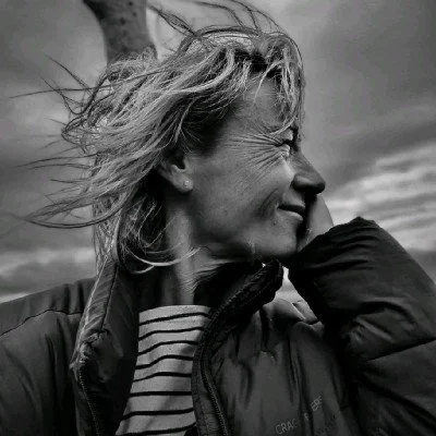 Side profile of a woman smiling outdoors with wind-blown hair, wearing a jacket and striped shirt, on a cloudy day.