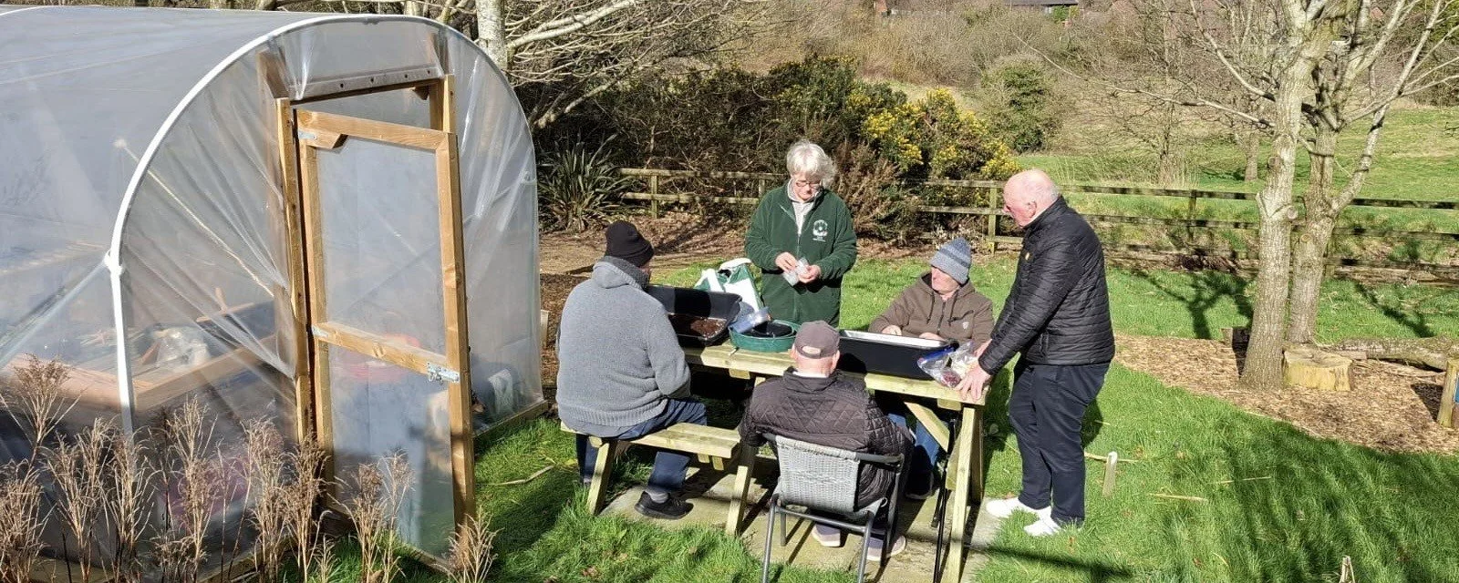 A group of five people are gathered outside on a grassy area around a picnic table, with a small greenhouse nearby. Some are seated, some standing, and they appear to be preparing or serving food on a sunny day.