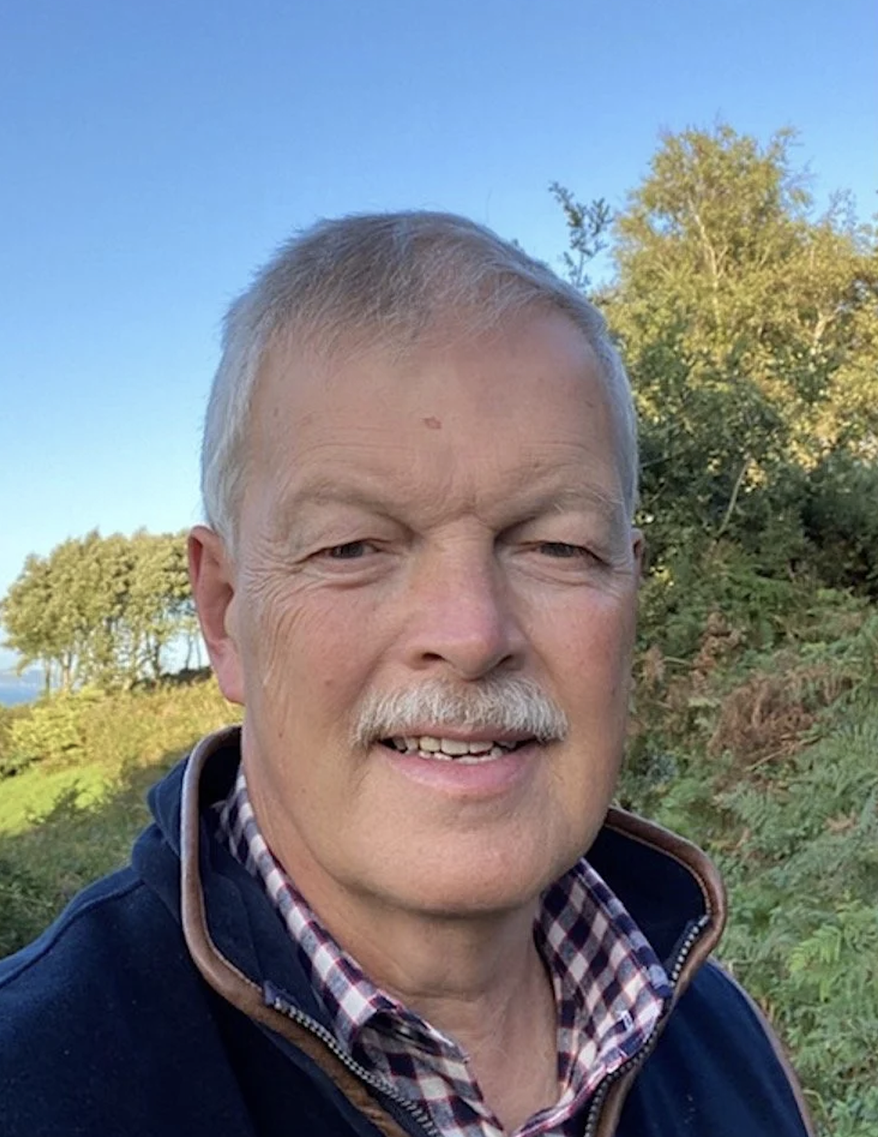 A man with short gray hair and a mustache smiling outdoors on a sunny day, with trees and blue sky in the background.