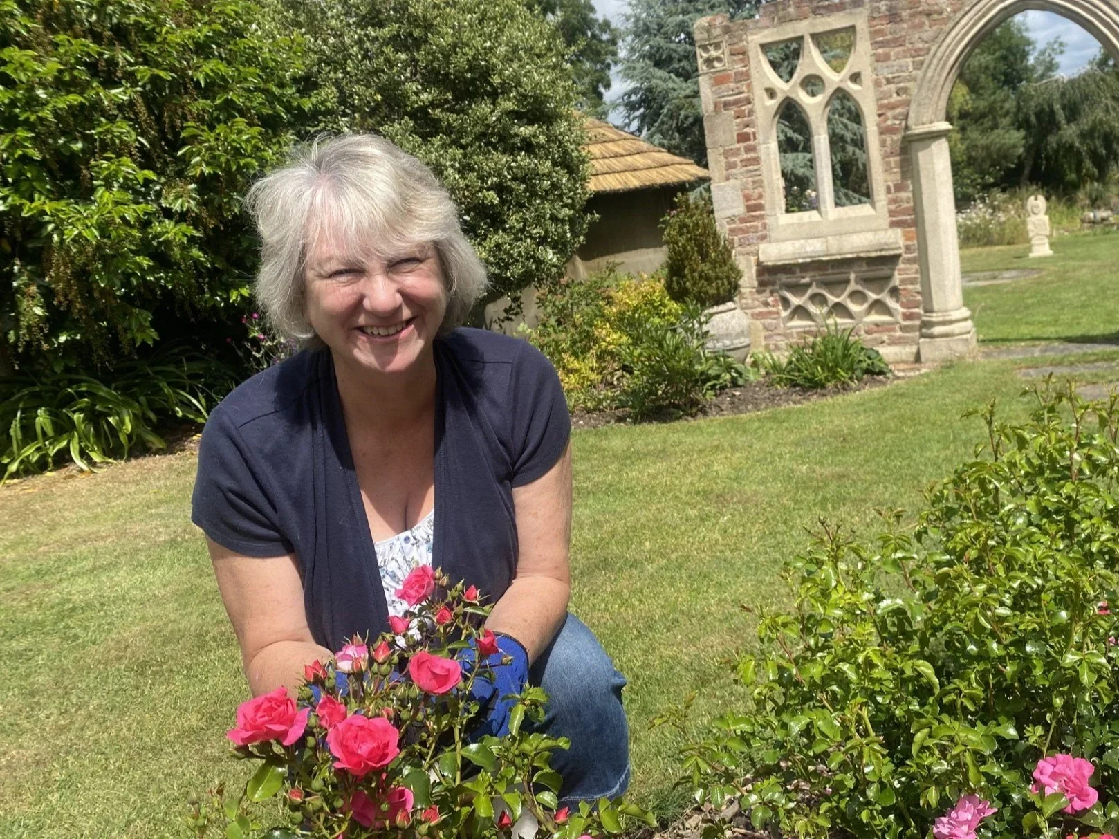 A woman with short gray hair smiling while crouching near pink flowers in a garden, with a stone and brick building and green bushes in the background.