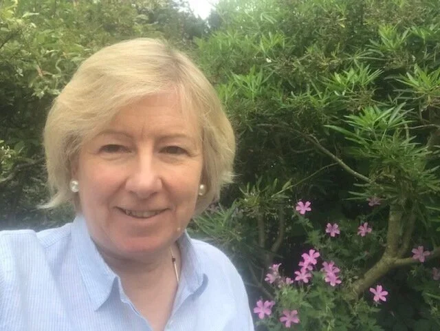 A woman with blonde hair smiling outdoors with green foliage and pink flowers in the background.