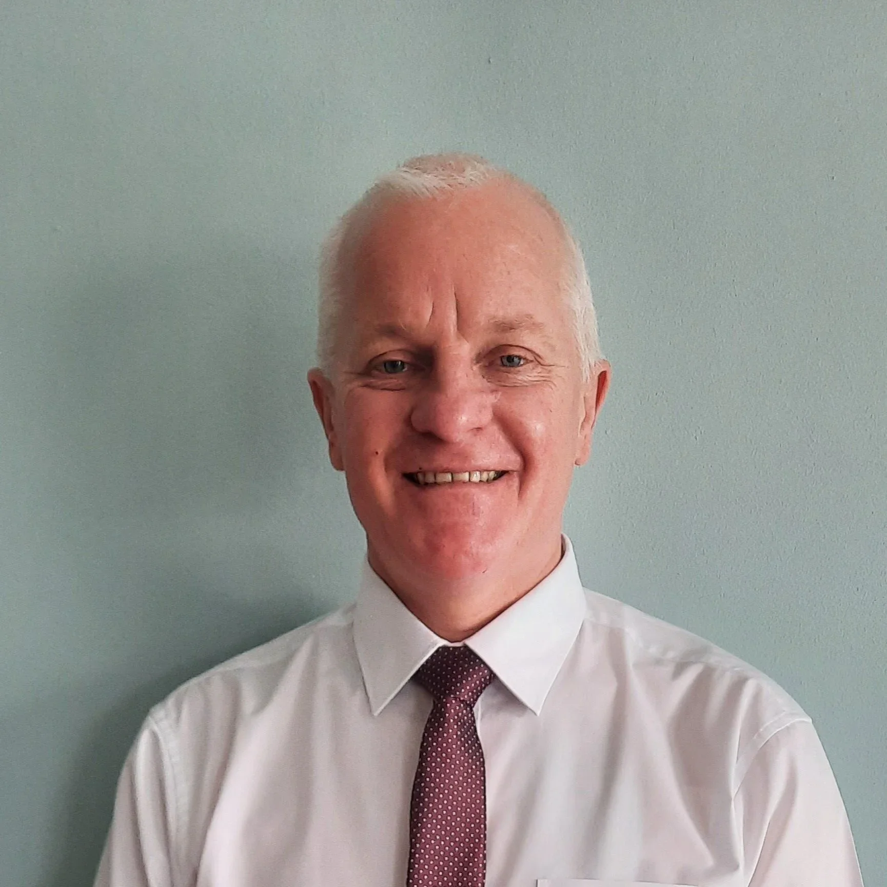A smiling man with gray hair in a white dress shirt and maroon tie standing against a light green wall.
